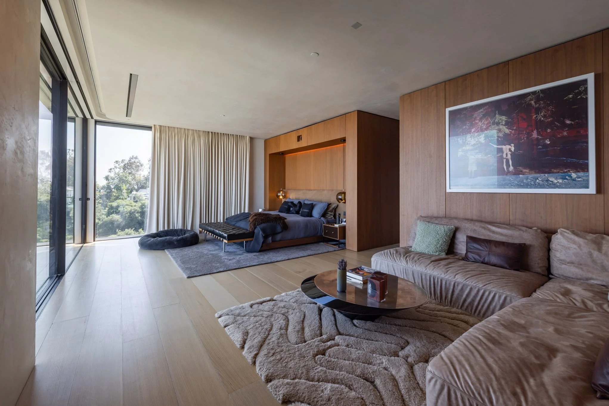 Modern hotel room with a large window, a bed with dark bedding, a gray rug, a beige sectional sofa, a wooden coffee table, and minimalistic decor, including a framed picture on the wooden wall.