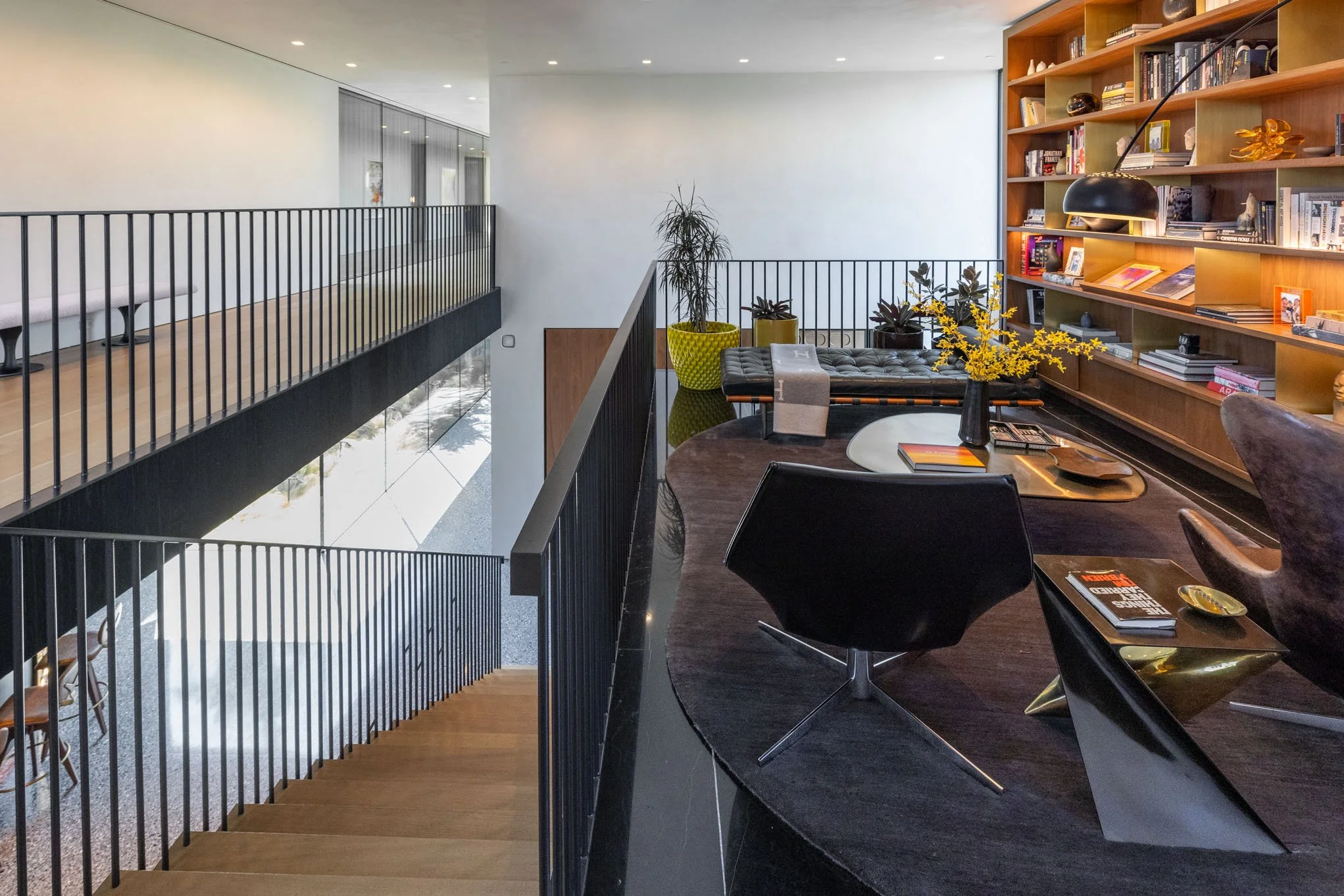 Interior view of a modern house with a second-floor balcony overlooking a living space. The space has a dark wood table, black chairs, a large built-in wooden bookshelf with books and decor, and a few potted plants.