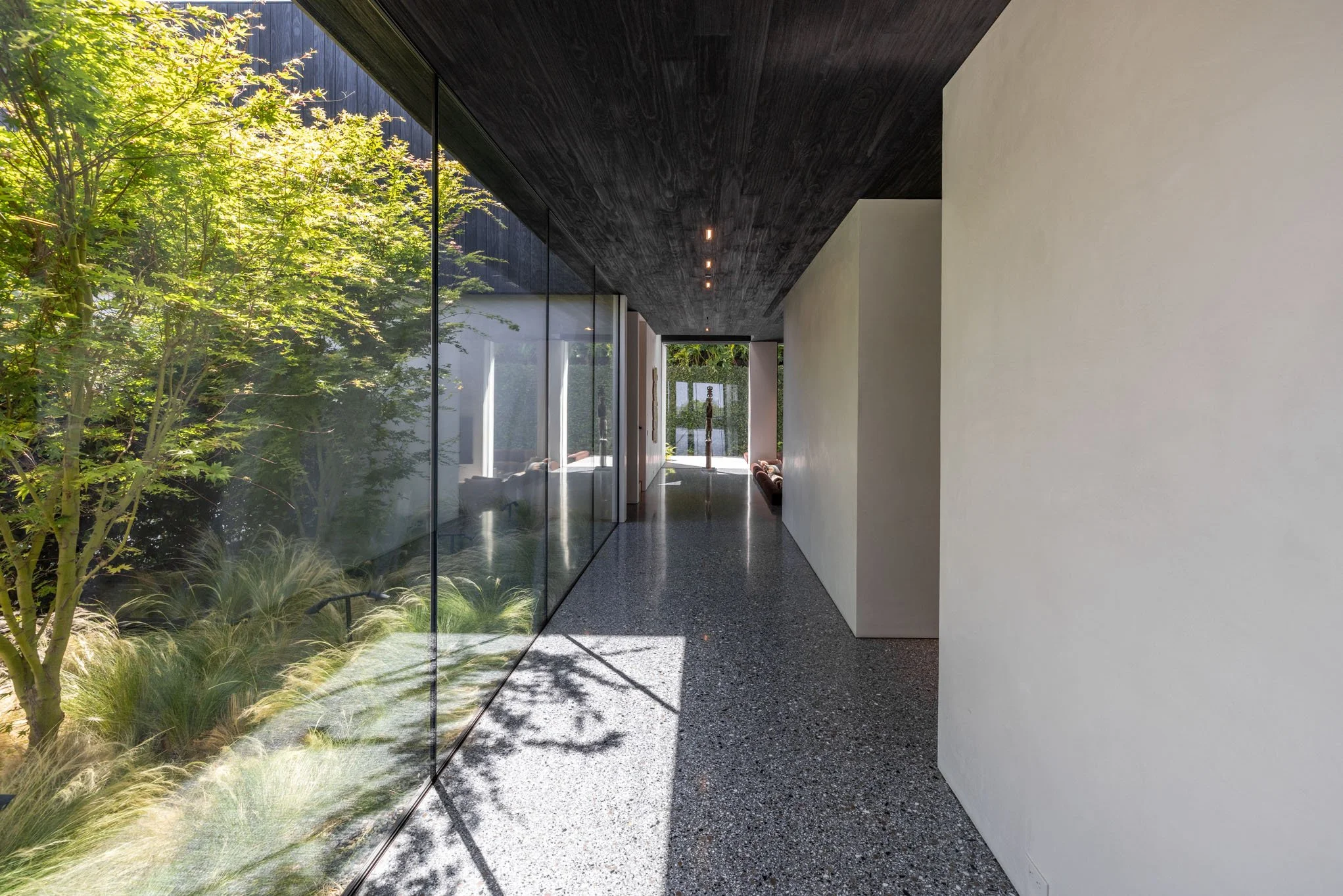 Modern interior hallway with large glass wall overlooking a garden, black ceiling with recessed lighting, white and beige walls, and terrazzo flooring.