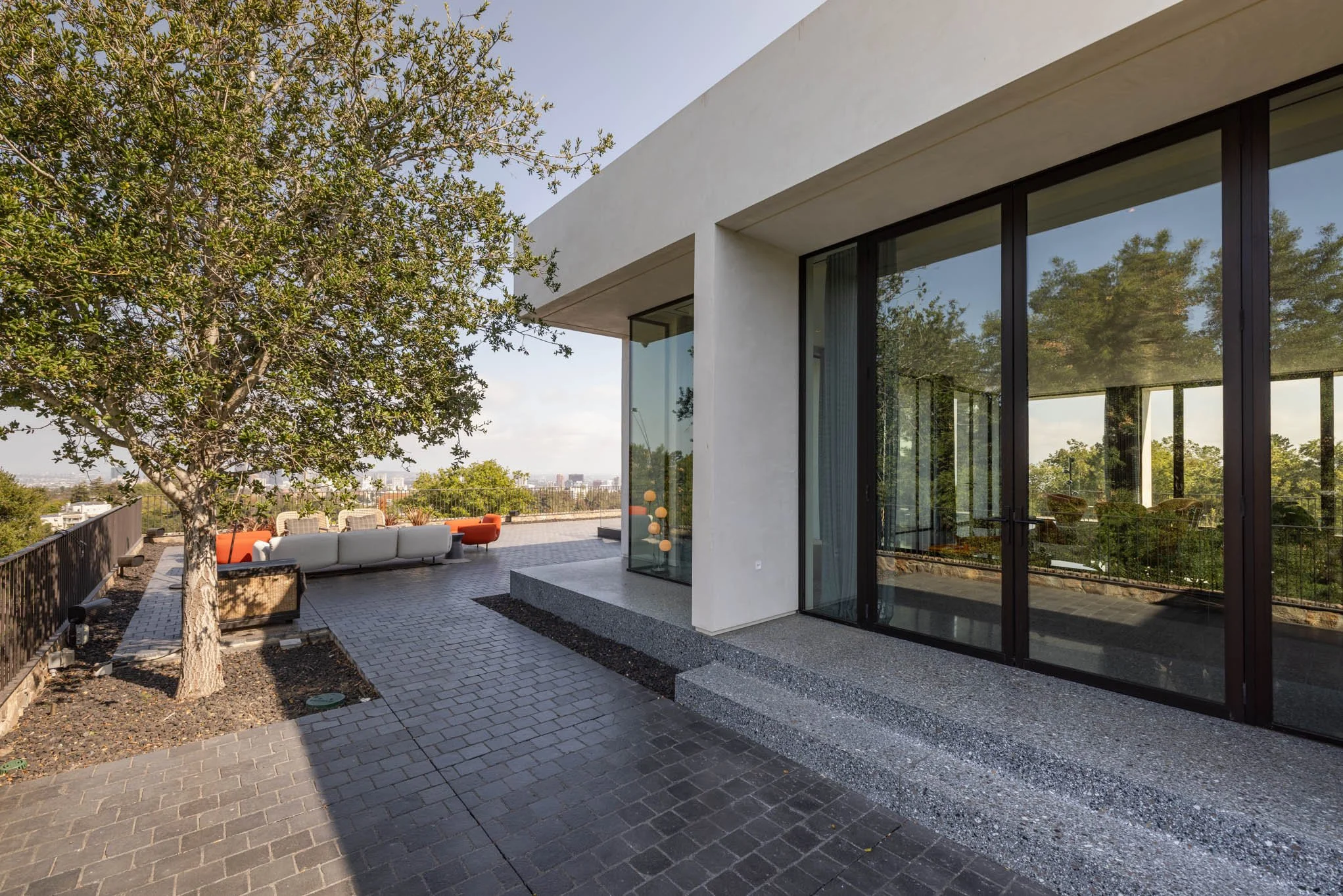 Modern building exterior with large glass sliding doors, outdoor seating area, tree, and cityscape view.