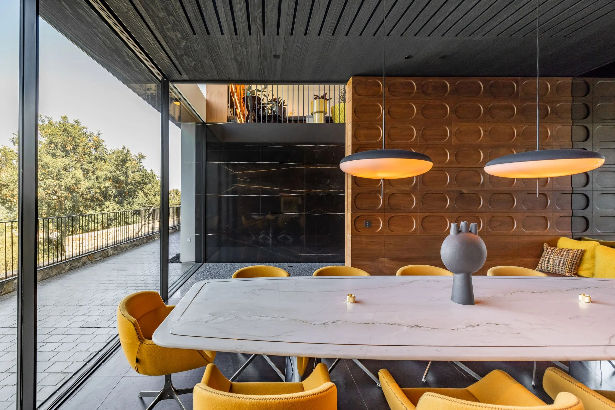 Modern dining room with a white marble table, yellow chairs, two hanging pendant lights, and a dark wooden accent wall.
