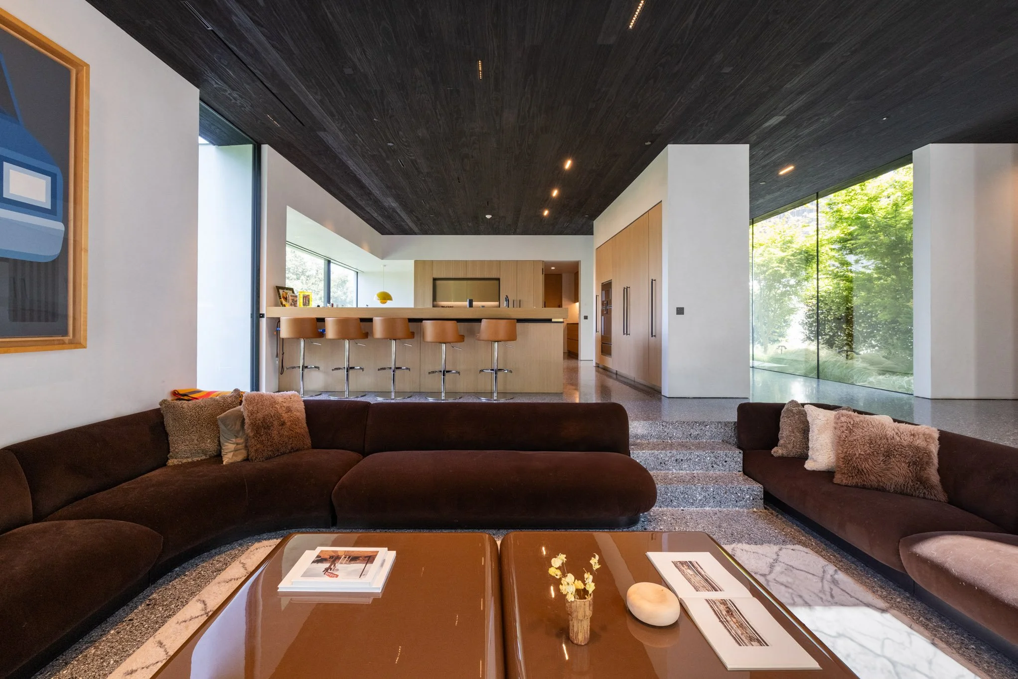 Modern living room with brown couches, a wooden coffee table, and decorative pillows. In the background, an open kitchen with beige bar stools and a black countertop, large windows with greenery outside, and a dark wood ceiling.