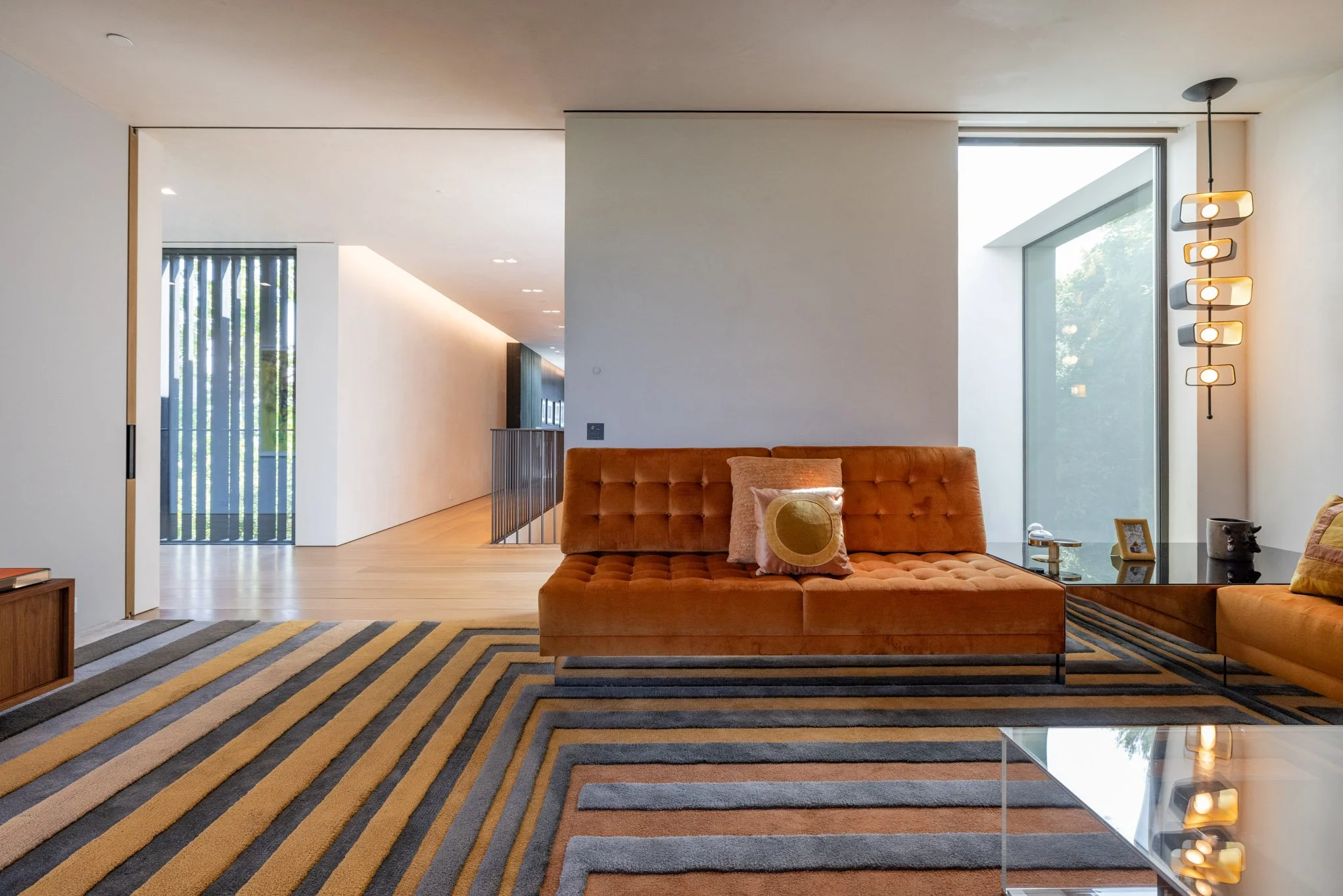 Modern living room with an orange tufted sofa, amethyst pillows, a glass side table with decorative items, and a geometric light fixture, large window, and a patterned yellow, gray, and black rug.