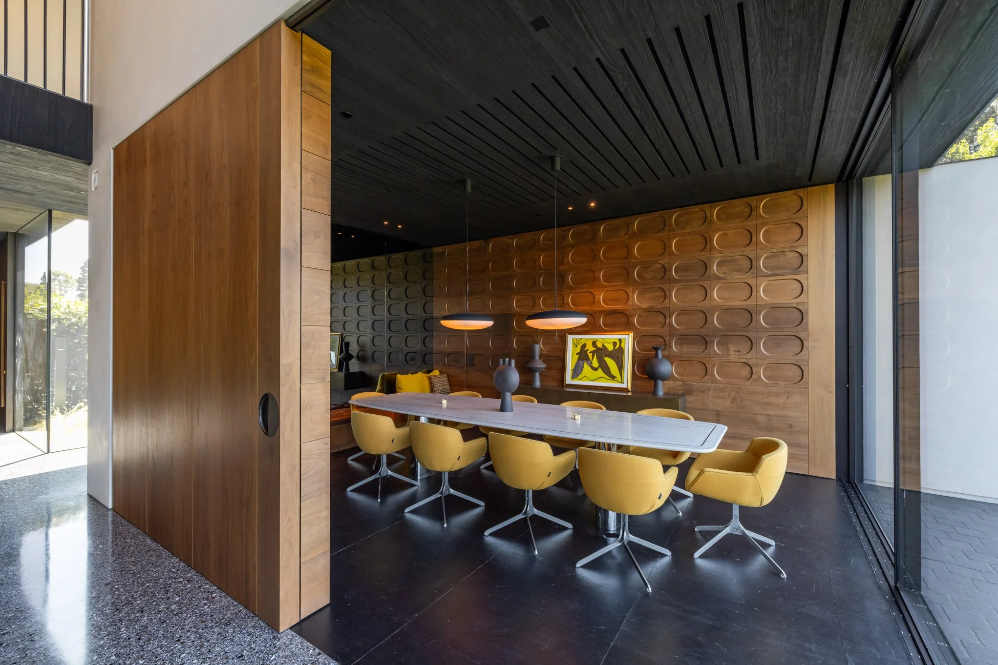 Modern dining room with a large white table, yellow chairs, dark ceiling with pendant lights, a wooden wall with decorative patterns, and large windows with a view outside.