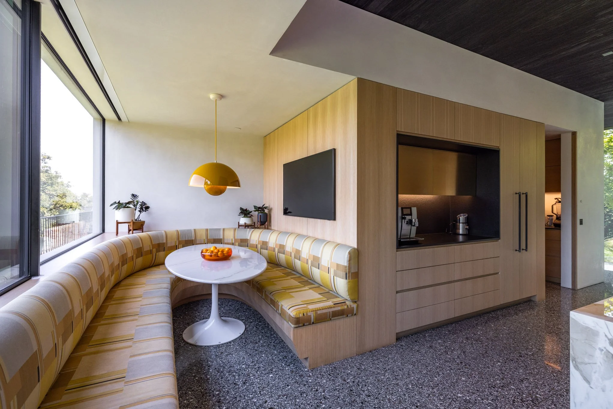 Modern dining area with a curved yellow checked banquette, white round table, hanging yellow pendant light, large window, and wood-paneled wall with a black TV