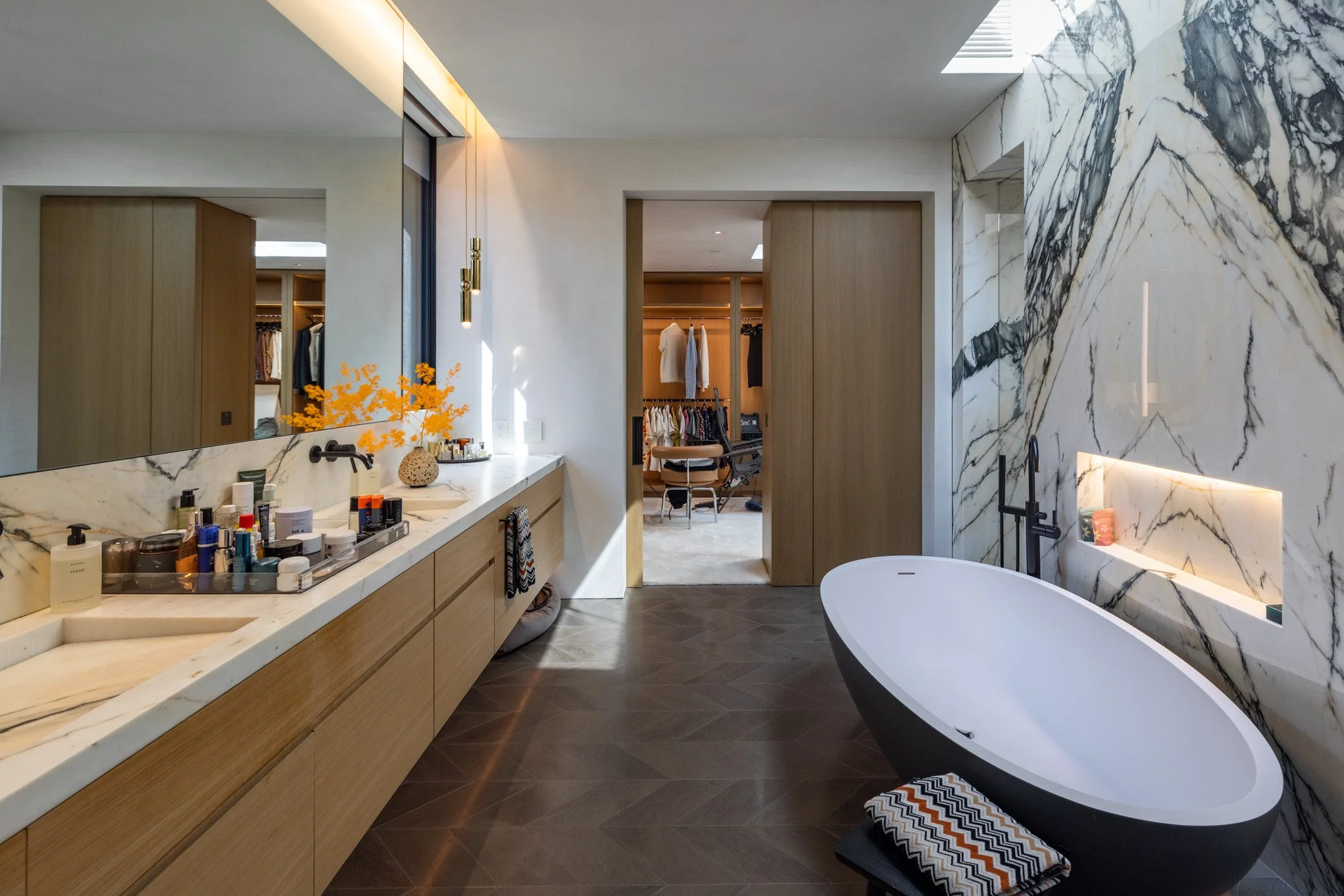 Modern bathroom with a black oval bathtub, a large marble wall, a long countertop with various toiletries, and a doorway leading to a walk-in closet with hanging clothes.