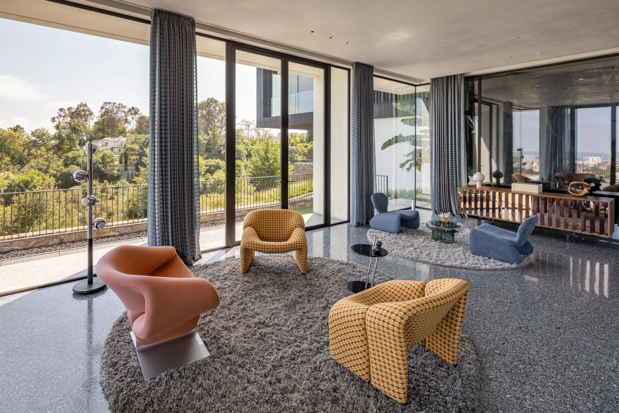 Modern living room with large floor-to-ceiling windows, gray curtains, a gray terrazzo floor, a shaggy gray area rug, and colorful contemporary chairs including a pink one, yellow patterned chairs, and blue lounge chairs. Outside, green trees are vis