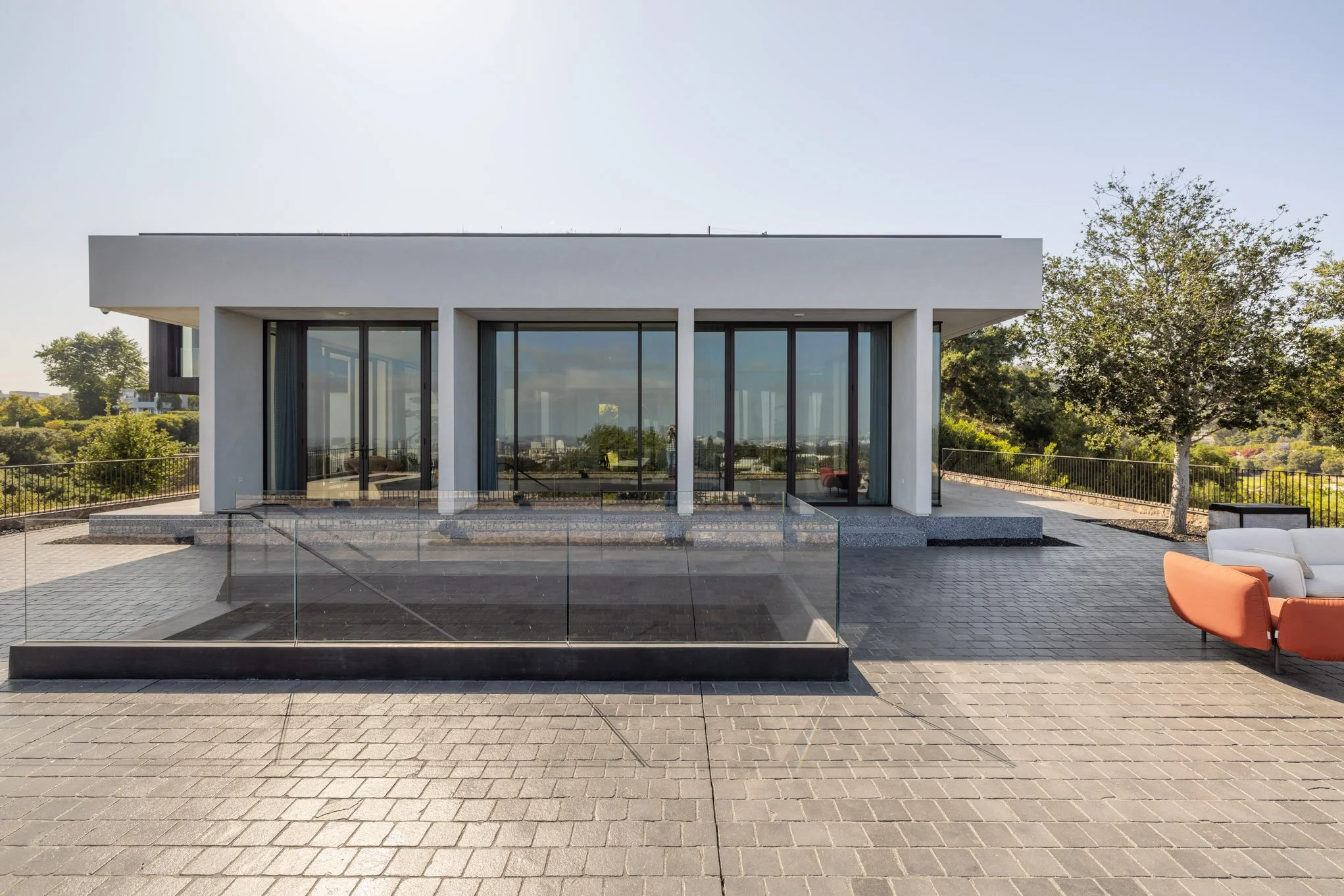 Modern white building with large glass doors and windows, outdoor seating with orange and white chairs, on a paved patio with a glass barrier, surrounded by trees and a landscape in the distance.