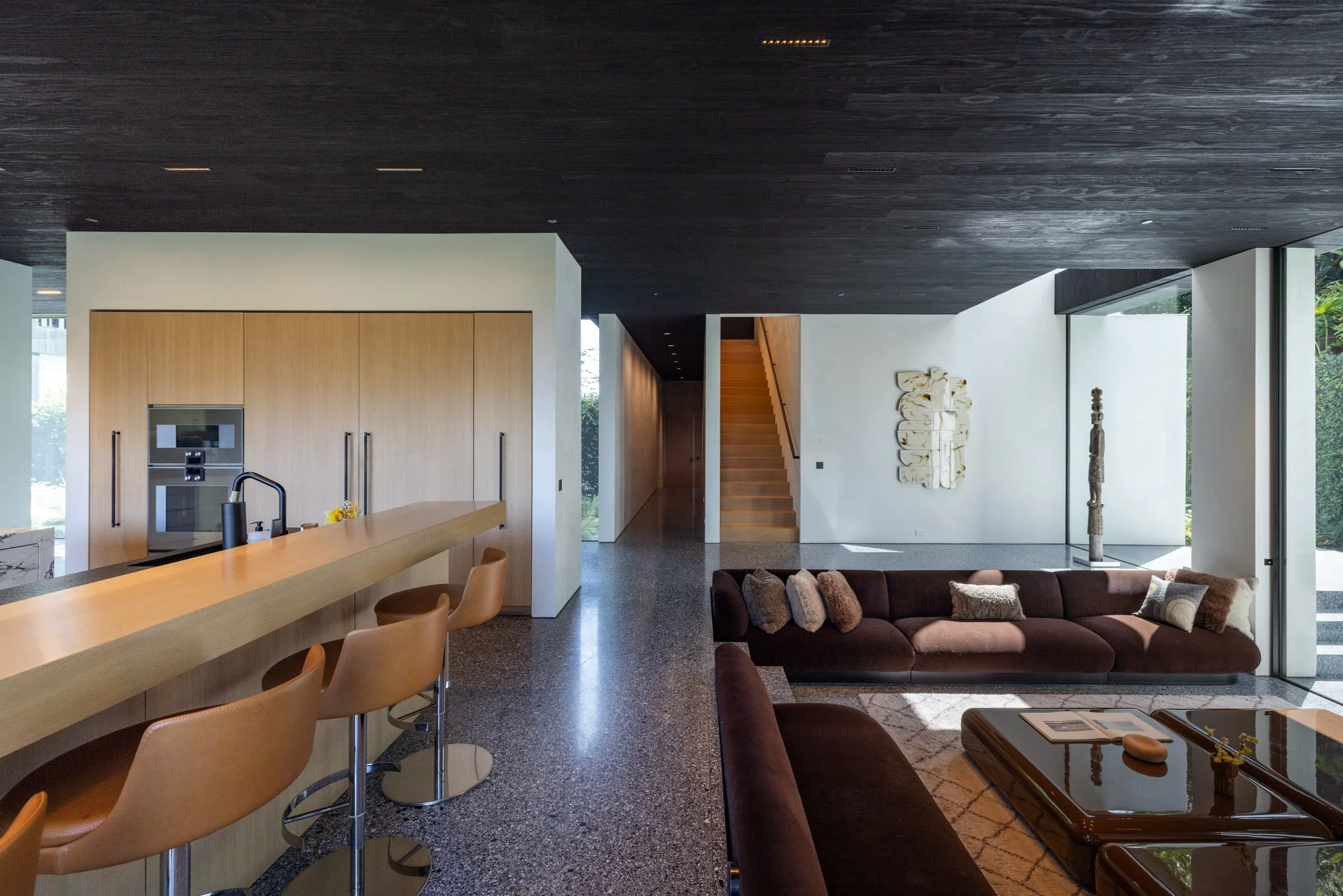 Modern open-concept living room and kitchen area with wooden accents, a staircase, and large windows allowing natural light.