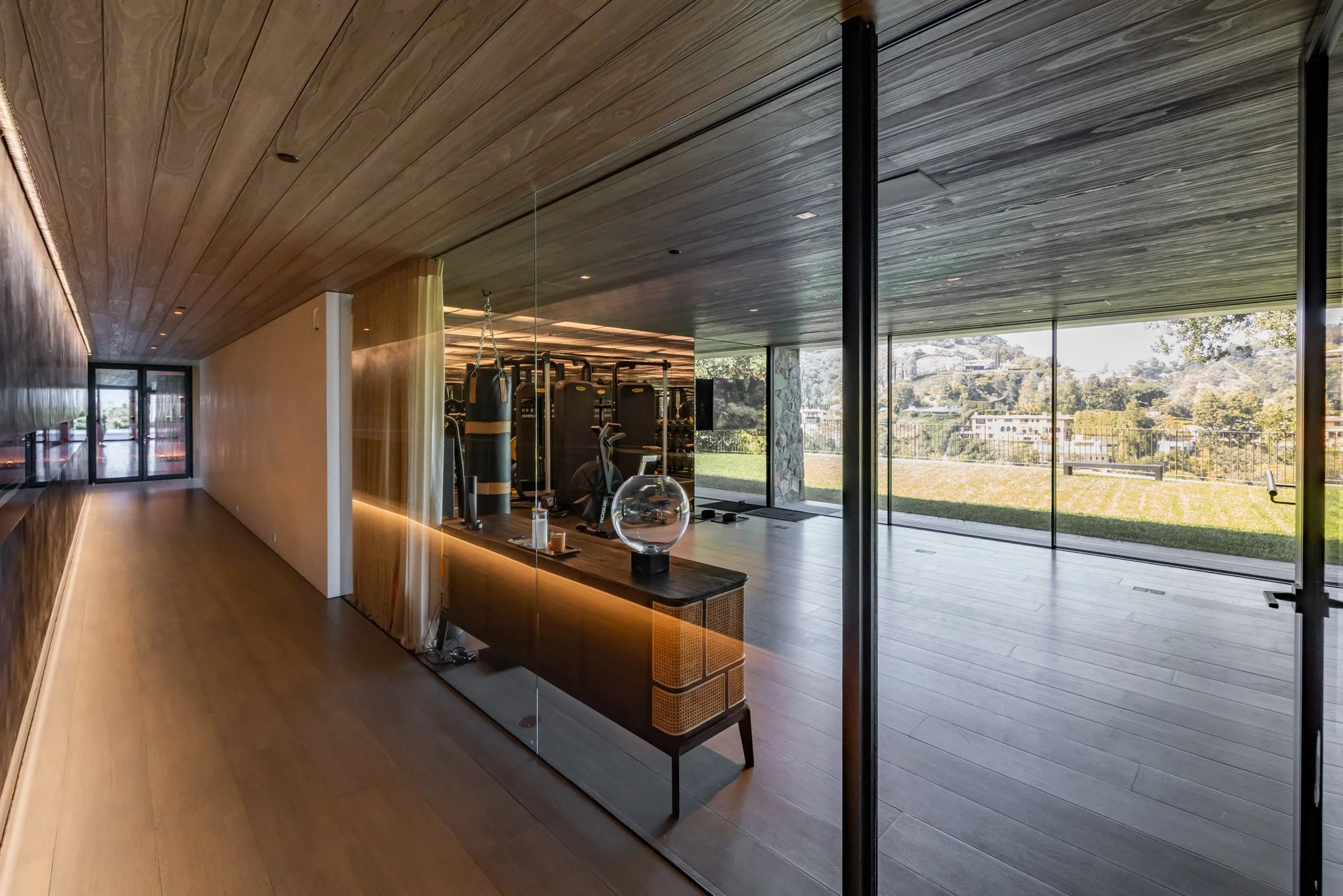 Modern gym with large glass walls overlooking a grassy outdoor area, featuring elliptical trainers, punching bags, and fitness equipment inside.