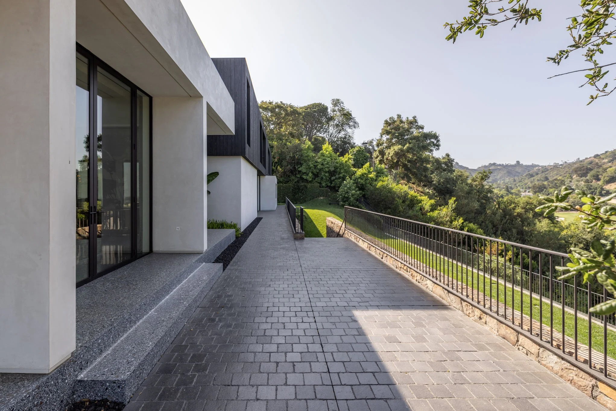 Modern house with large glass sliding doors, concrete and brick walkway, and a view of green trees and hills in the background.