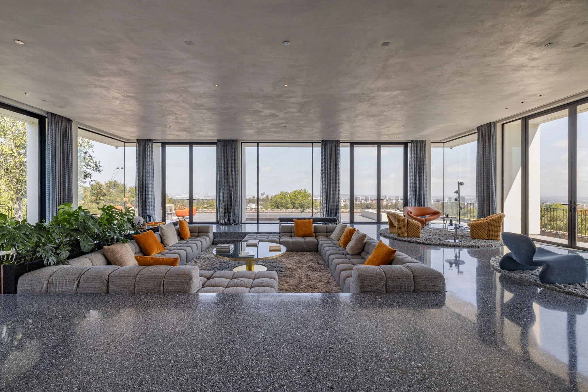 Modern living room with large floor-to-ceiling windows, gray sectional sofa with orange and beige pillows, a round coffee table, and various seating areas on white and gray rugs, overlooking a cityscape on a sunny day.