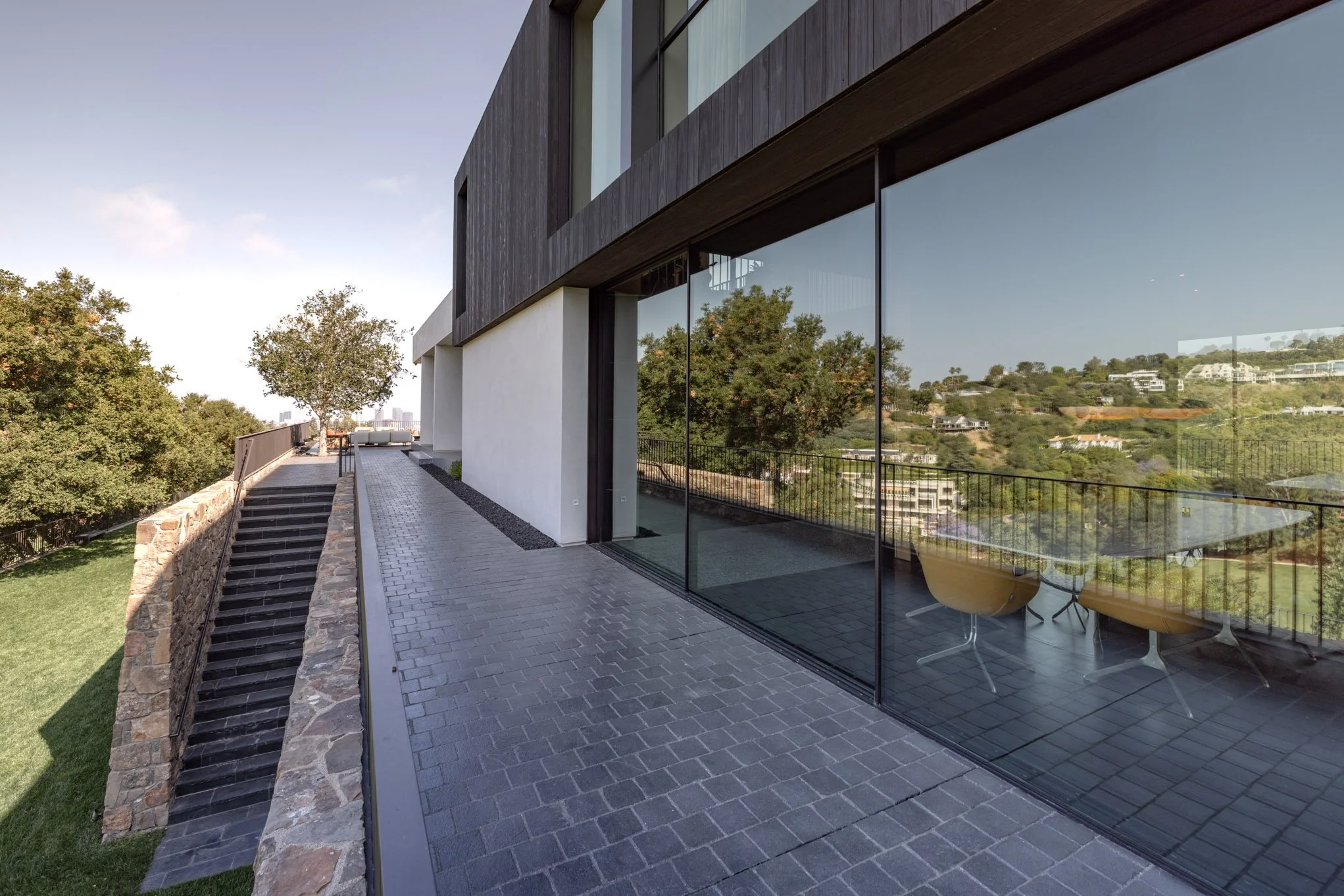 Modern building with large glass windows reflecting trees and hillside, outdoor paved walkway, stone staircase, and patio furniture inside