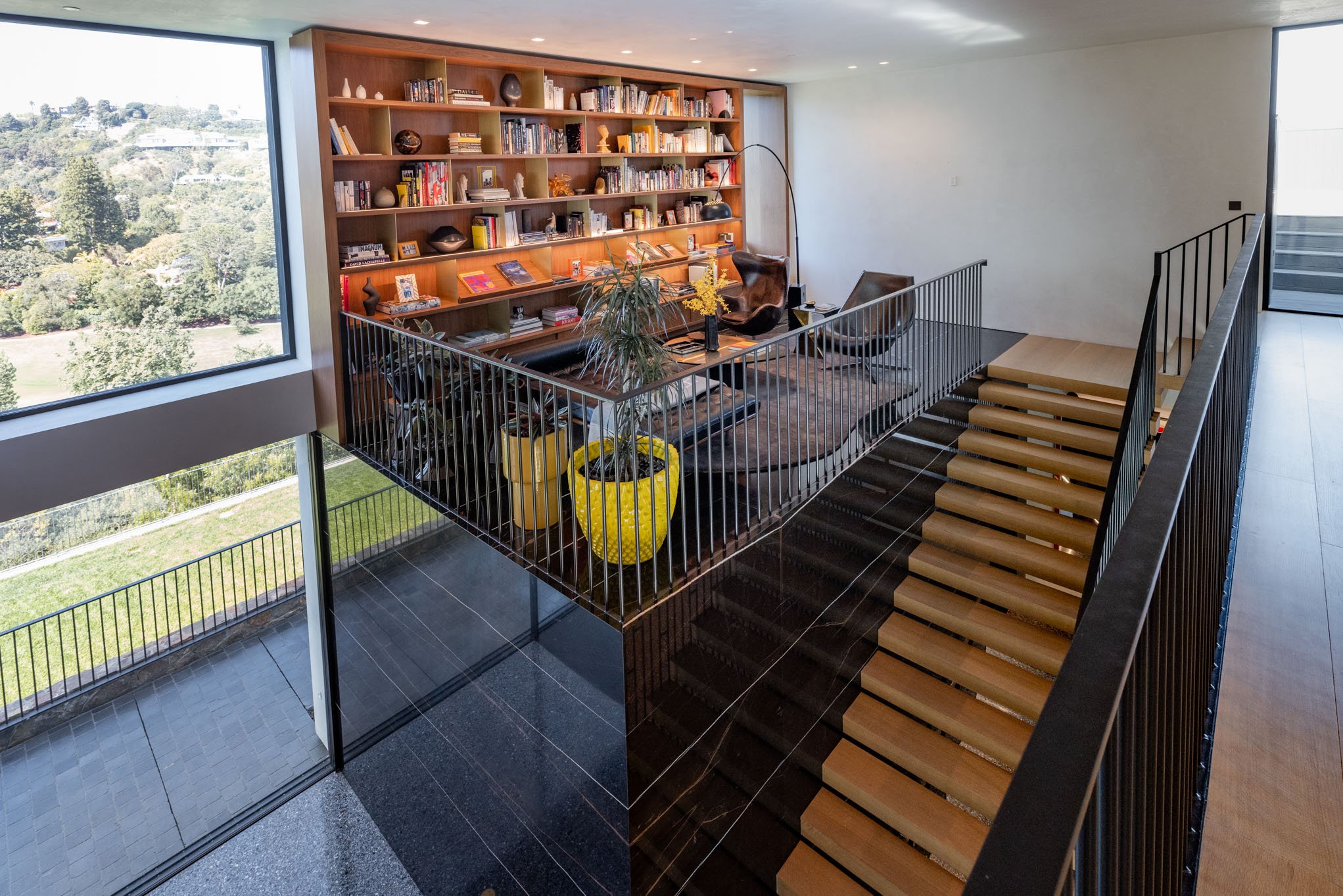 Interior view of a modern upper-level living space with a large wooden bookshelf, two brown chairs, a coffee table, and potted plants, overlooking a large window with a scenic outdoor landscape.