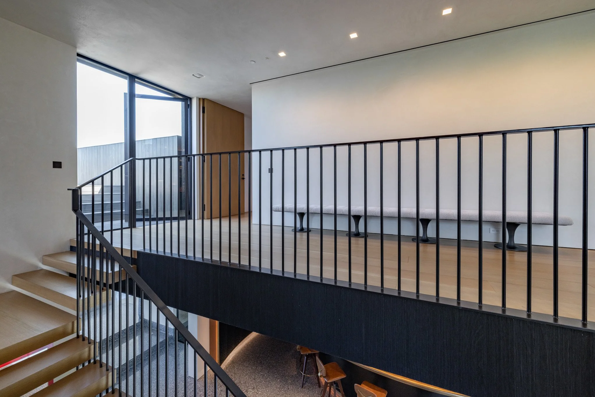 Interior view of a modern building with a staircase leading to a mezzanine level, featuring a black metal railing, wood flooring, a long cushioned bench against the white wall, and a large window with natural light.