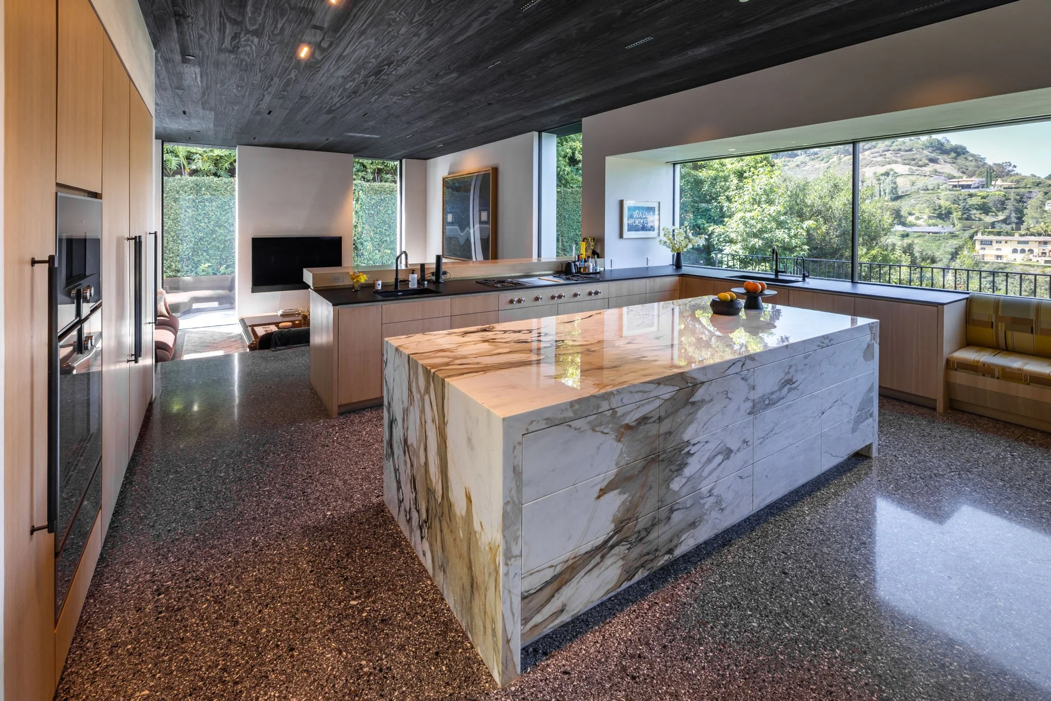 Modern kitchen with large marble island, light wood cabinets, black countertops, large windows overlooking green landscape, and a dark ceiling.