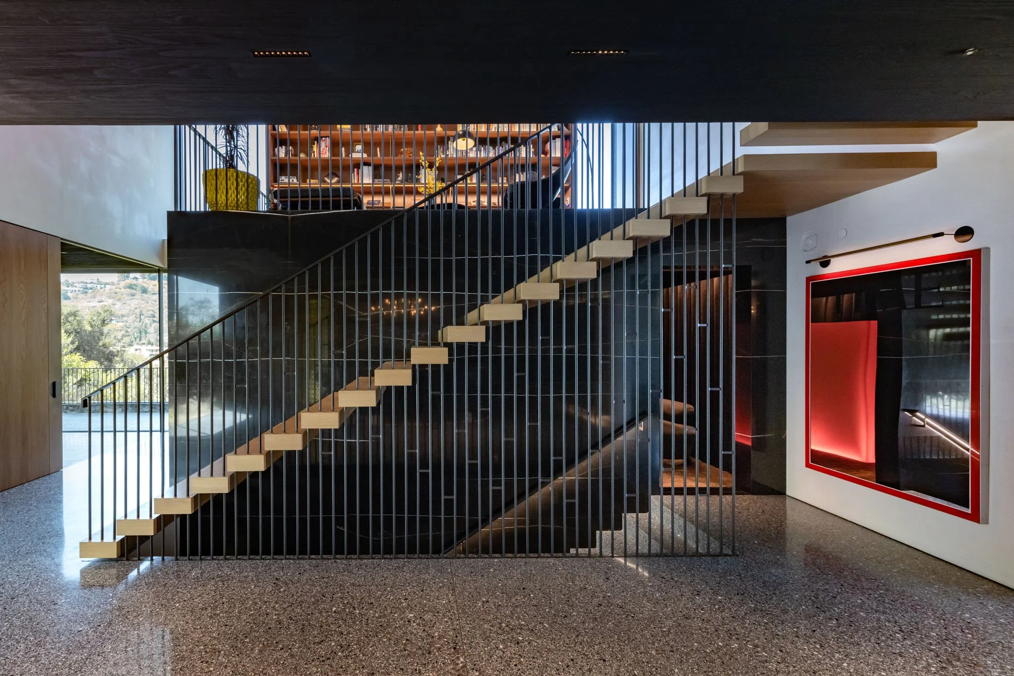 Modern interior with black staircase, wooden steps, metal railing, large window with scenic view, and a framed abstract art piece with red illumination on the white wall.