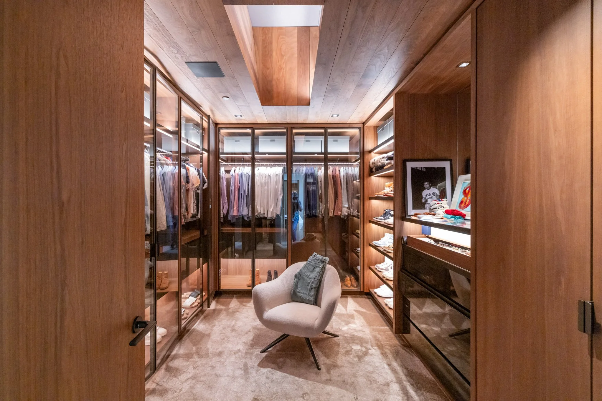 A walk-in closet with wooden walls, glass wardrobes filled with clothes, shelves with shoes, and a beige chair with a folded denim shirt on it.