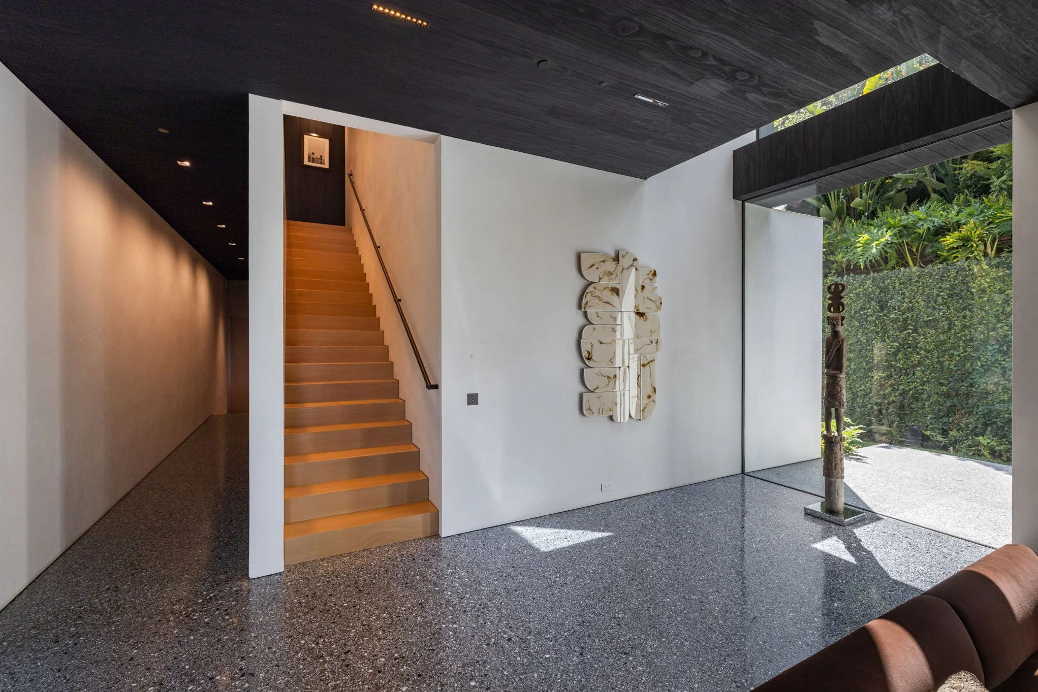 Modern interior with wooden staircase, abstract wall art, tribal sculpture, and large window with outdoor greenery.