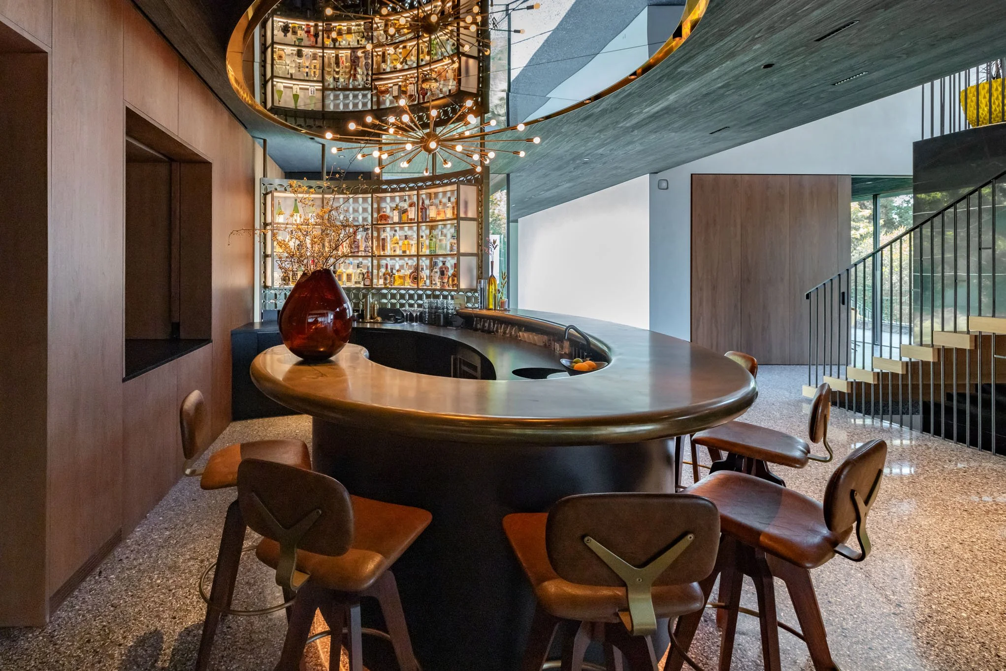 Modern indoor bar with a curved wooden counter, bar stools, a backlit display shelf with bottles, a decorative pendant light, large windows, and a staircase with black railings.