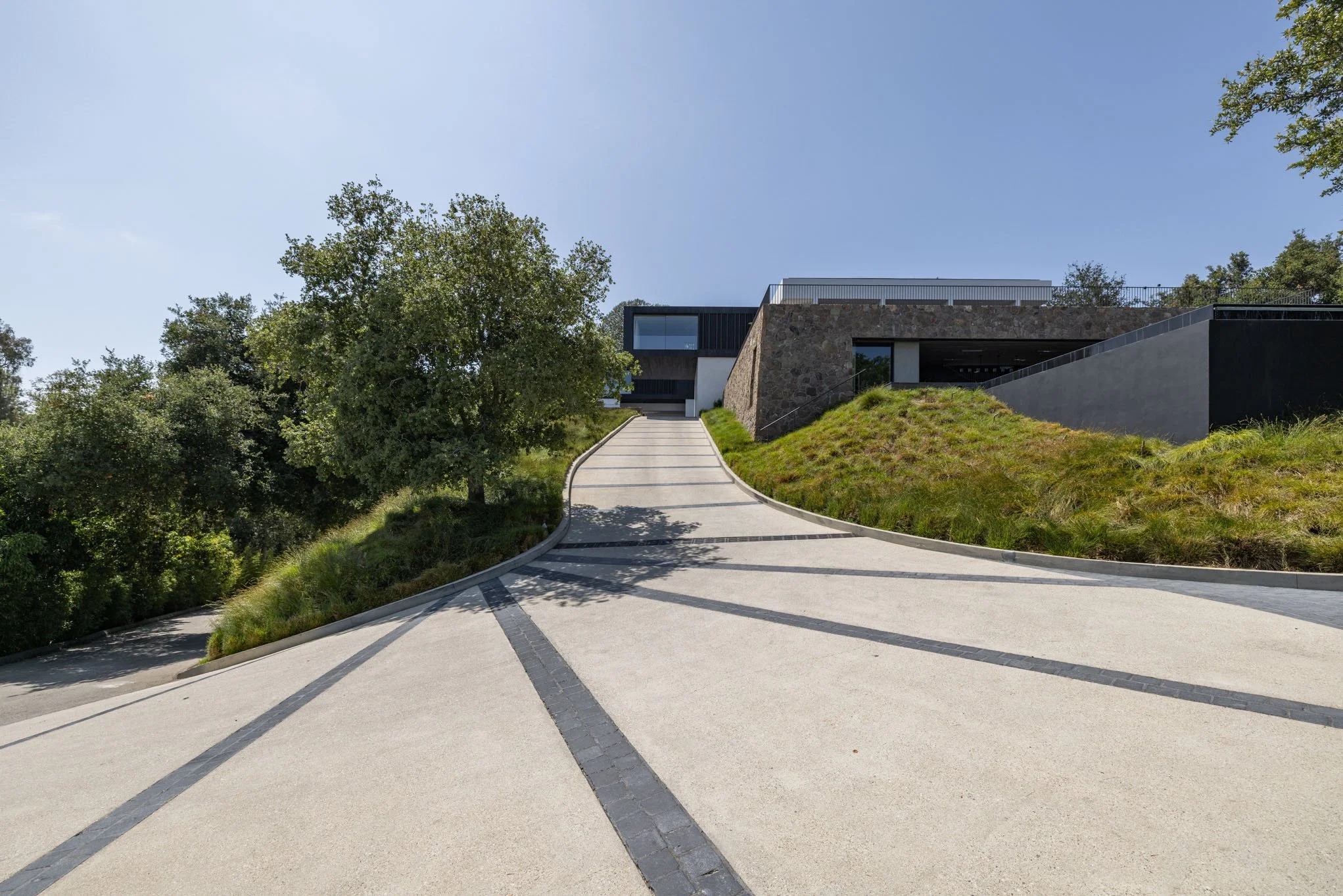 Modern house with a concrete driveway on a hill, surrounded by trees and grassy landscape, under a clear blue sky.