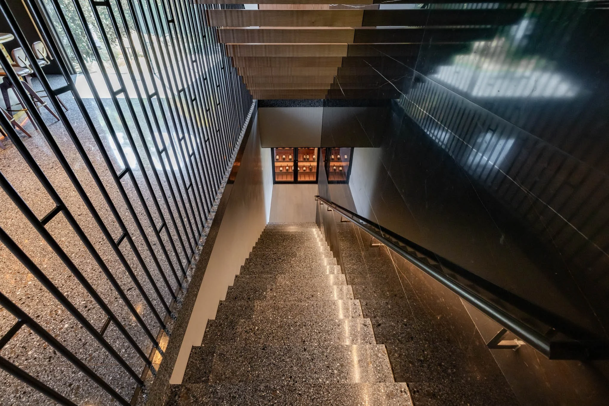 Looking down a staircase with a black granite railing on the right and black metal bars on the left, leading to a small landing with a glass door and a wine display.