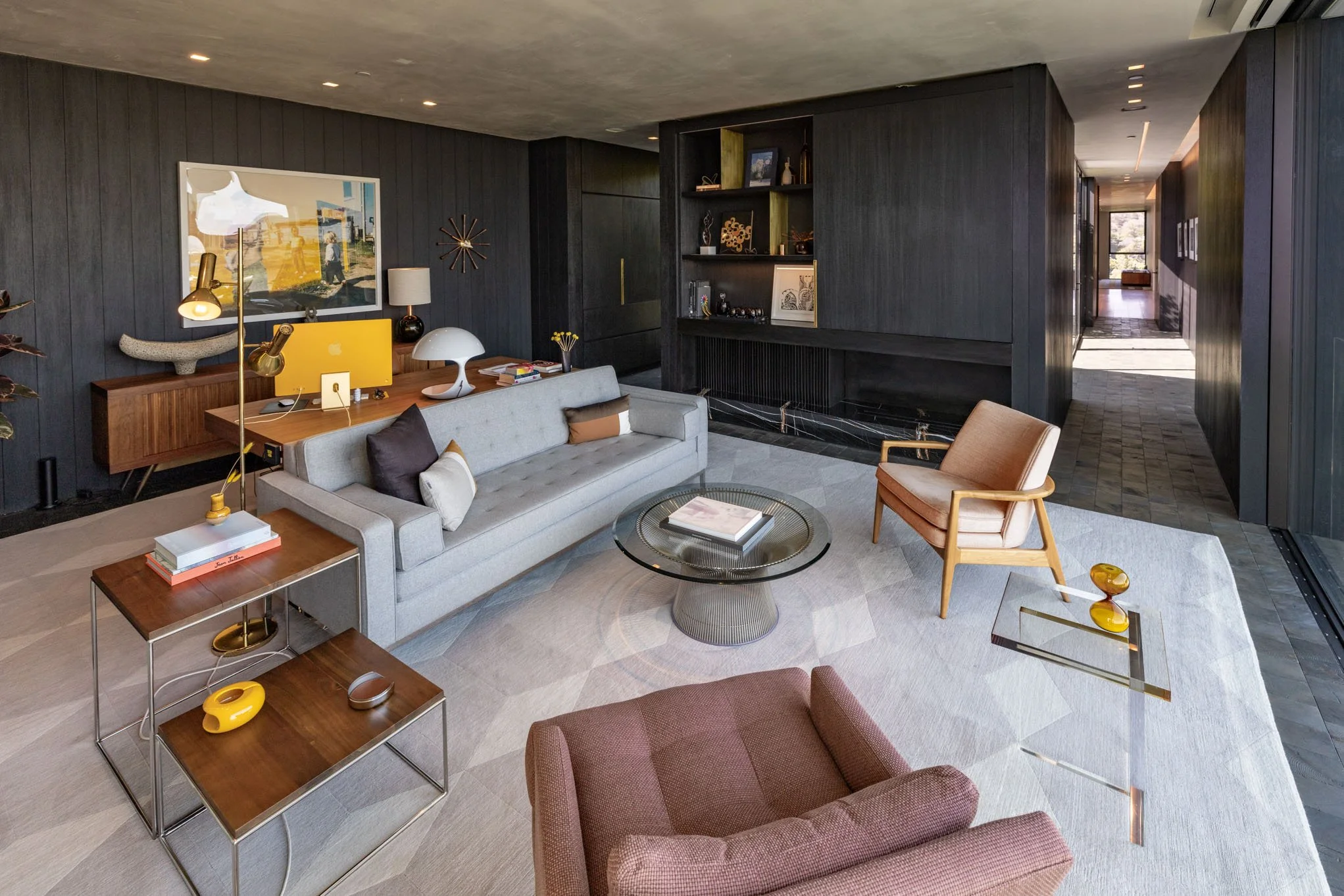 Modern living room with gray sofa, pink armchair, glass coffee table, dark wood wall panels, and decorative accents.