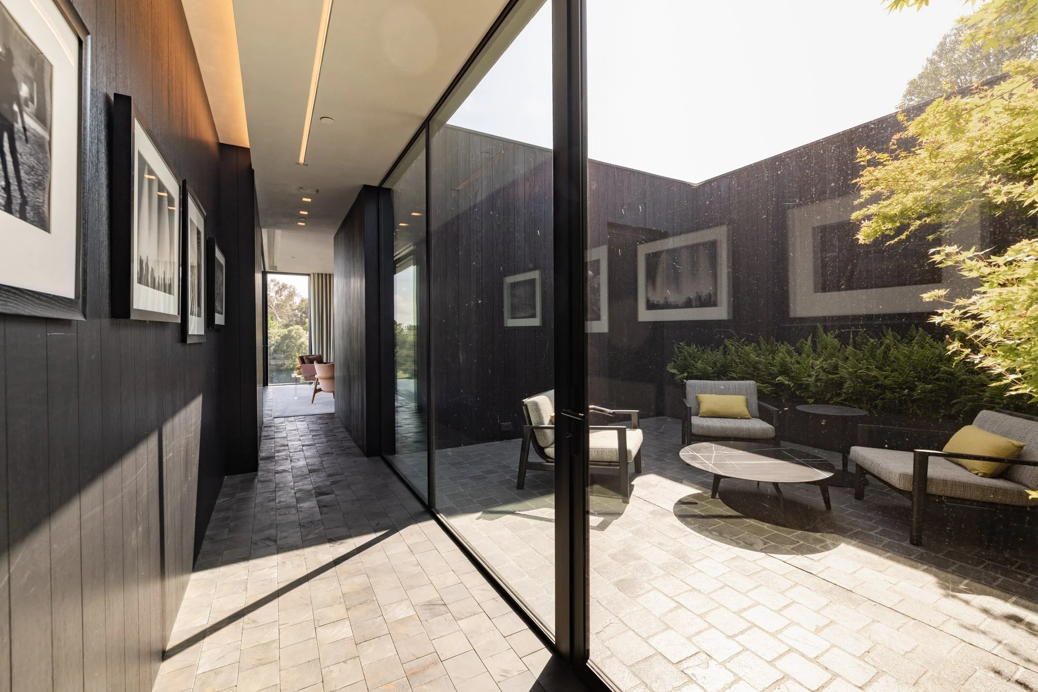 Interior view of a modern home with a hallway and an outdoor sitting area visible through a large glass wall. The hallway has artwork on the wall and leads to a space with a window showing trees outside.