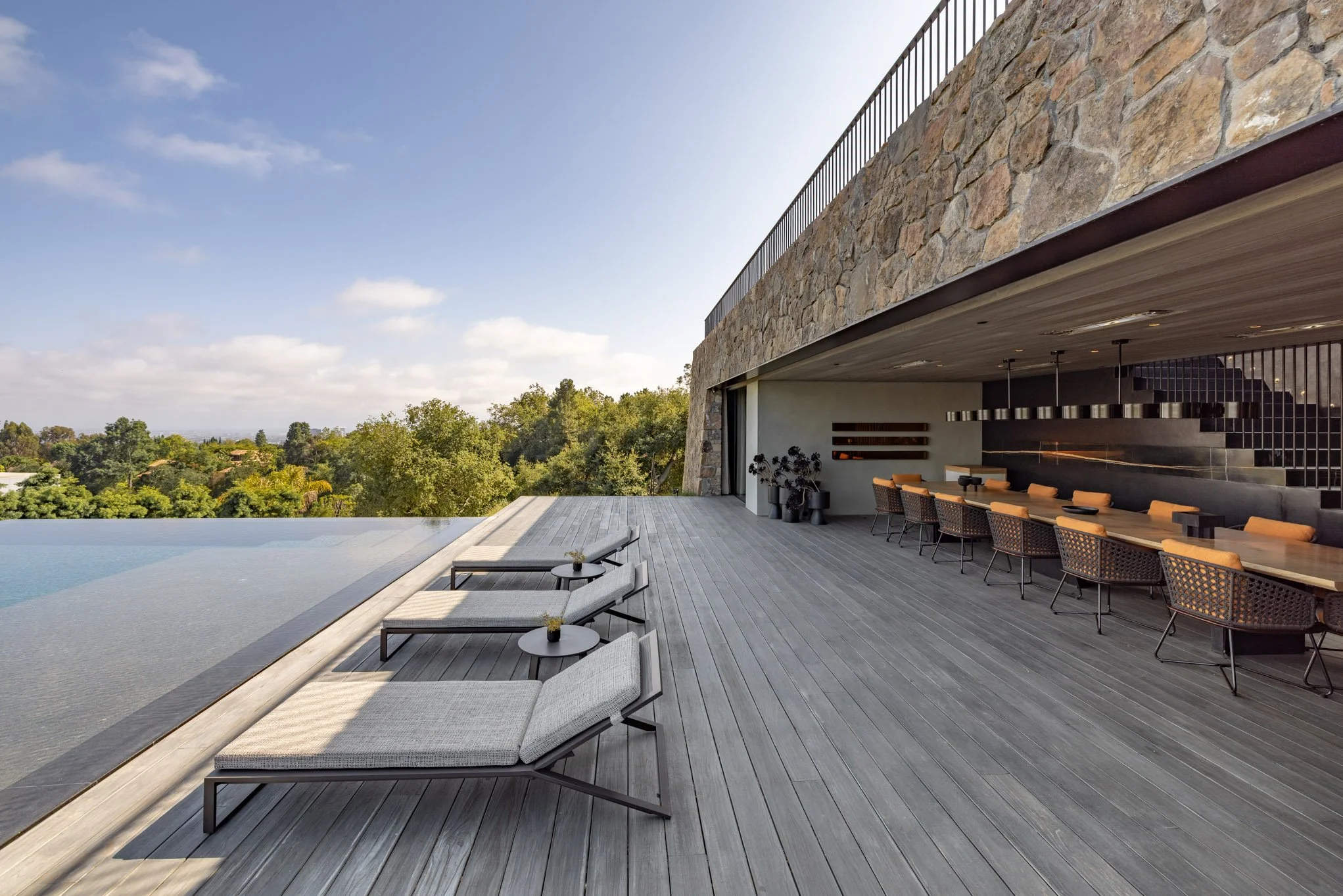 Modern outdoor infinity pool with lounge chairs and a seating area overlooking a lush green landscape under a partly cloudy sky.