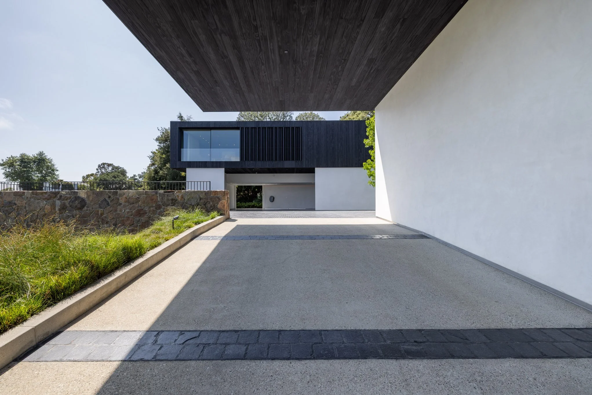 Modern house with white walls and black wooden accents, viewed from a driveway or parking area, with clear sky and some greenery in the background.