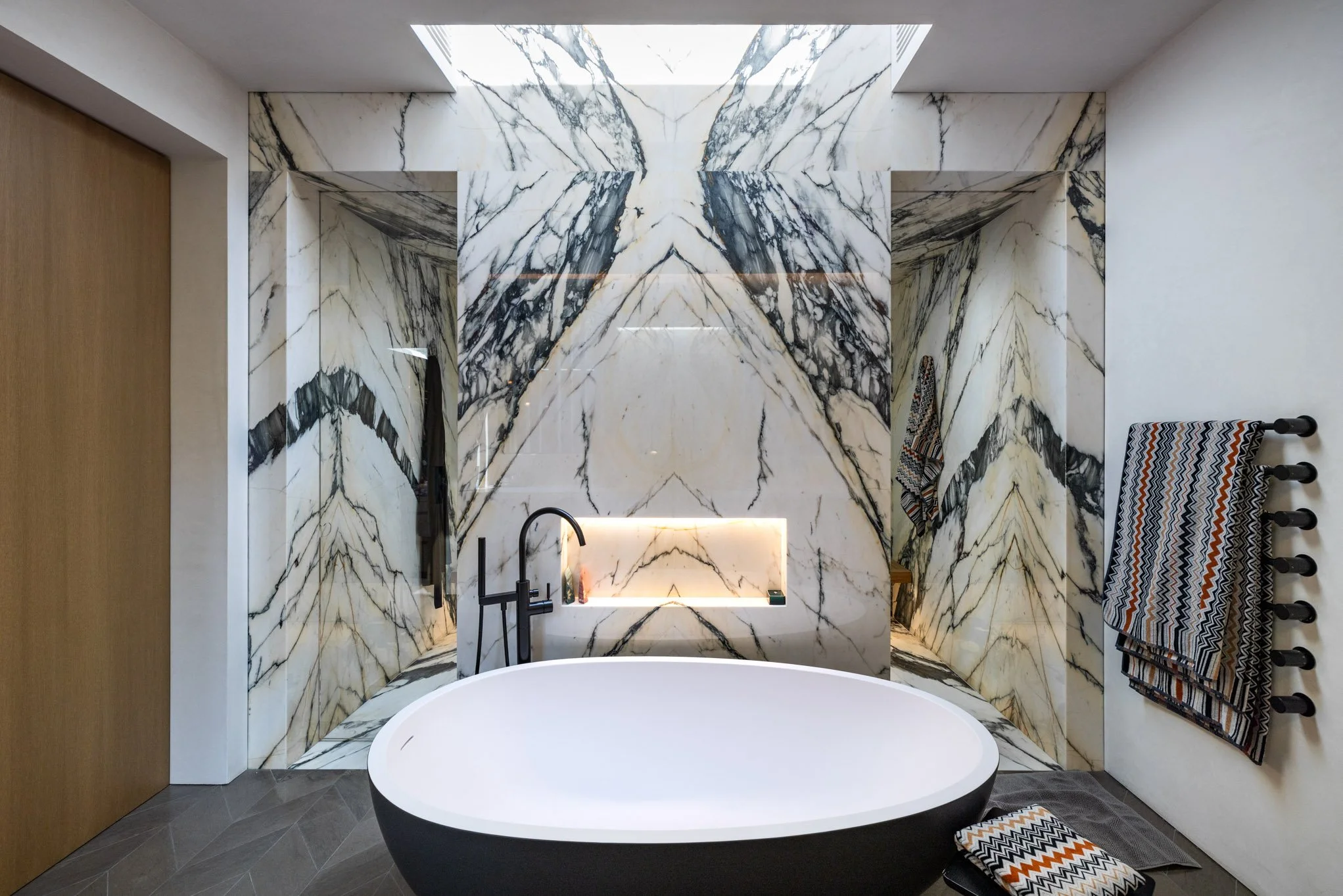 Modern bathroom with a black and white oval freestanding tub, surrounded by marble with black veining, complimented by a black faucet and towel racks, with striped towels and a small patterned bath mat.