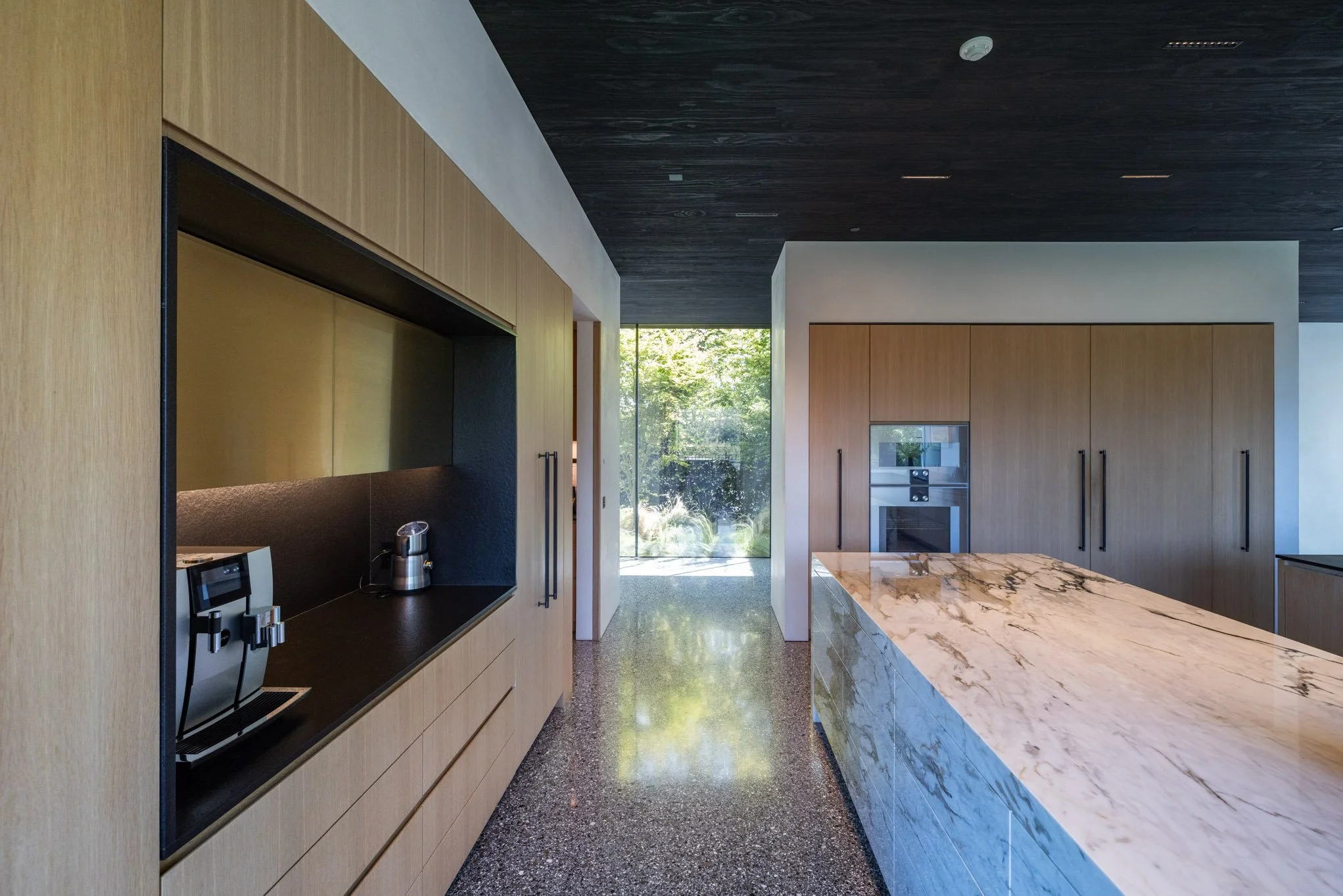 Modern kitchen with wooden cabinets, marble island, built-in oven, coffee machine, and large window with greenery outside.