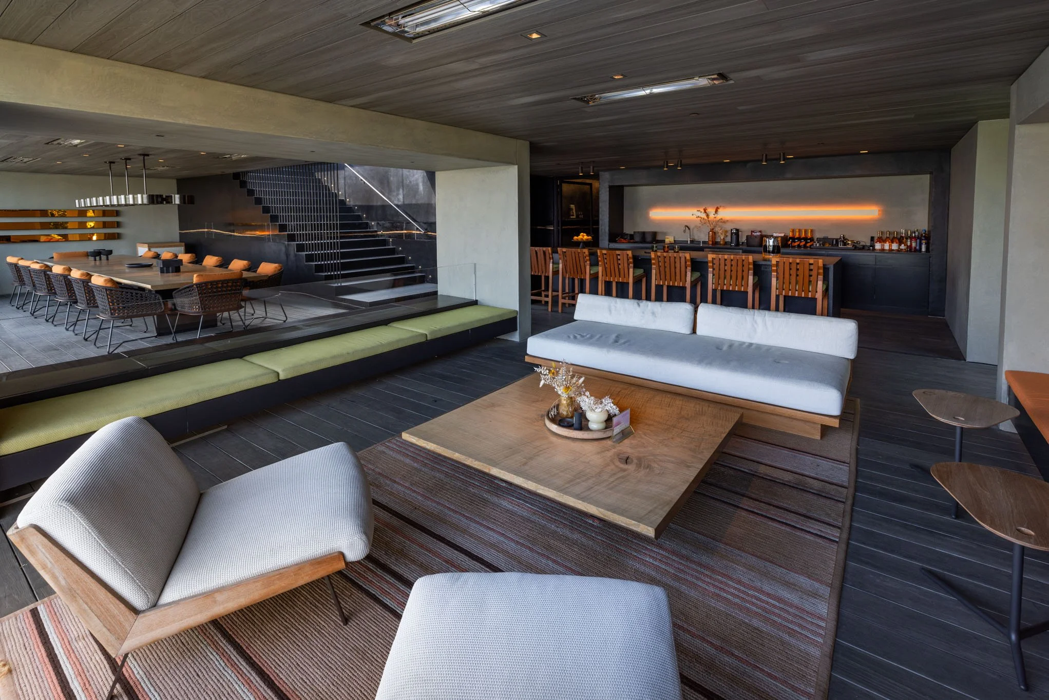 Modern, open-concept living and dining area with contemporary furniture, a large wooden coffee table, a white sofa, beige lounge chair, and a bar area with wooden chairs in a dimly lit, stylish interior.