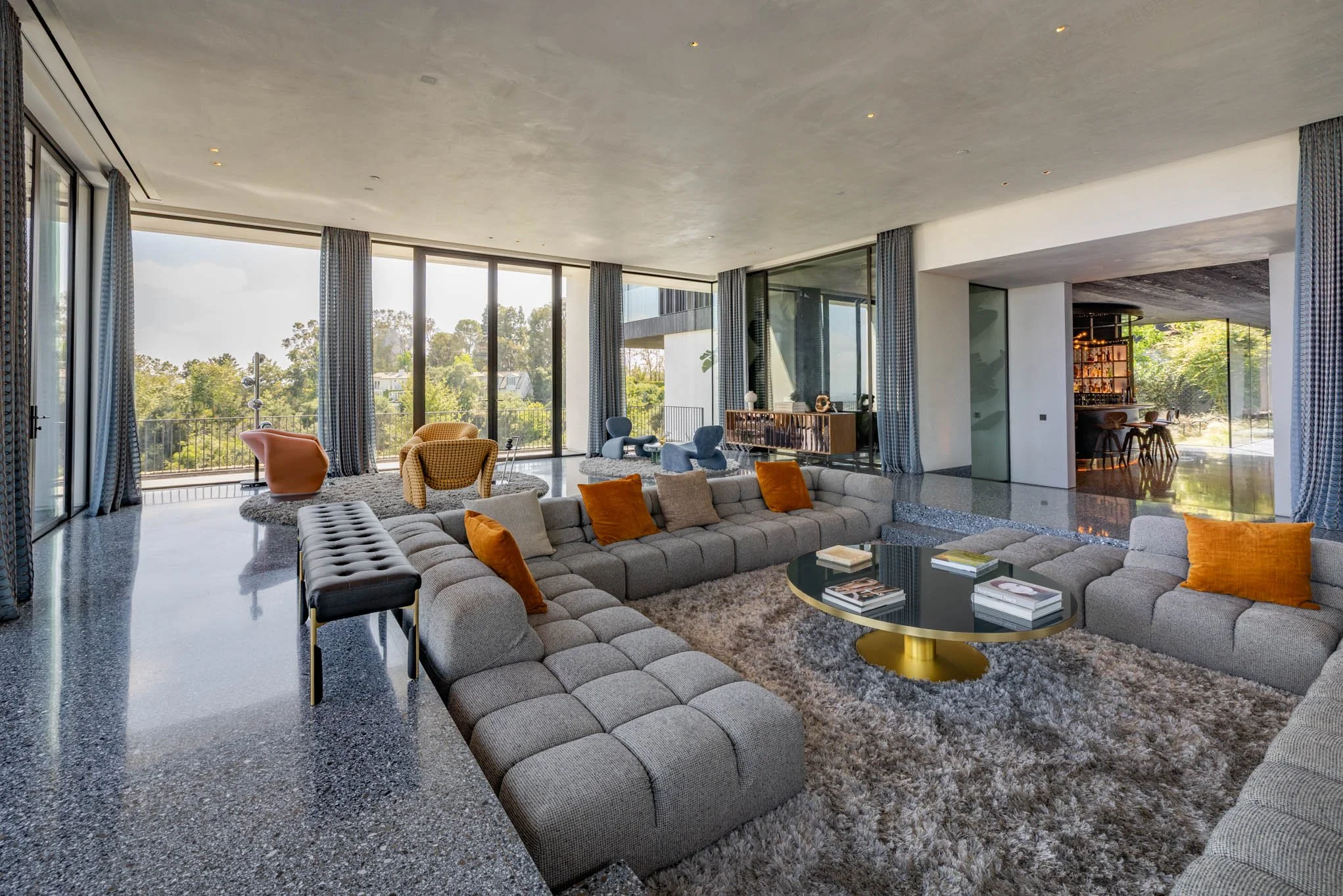 Spacious modern living room with large floor-to-ceiling windows, patterned curtains, gray sectional sofa with orange and beige pillows, shaggy rug, round black and gold coffee table with books, and colorful armchairs, overlooking a green outdoor land