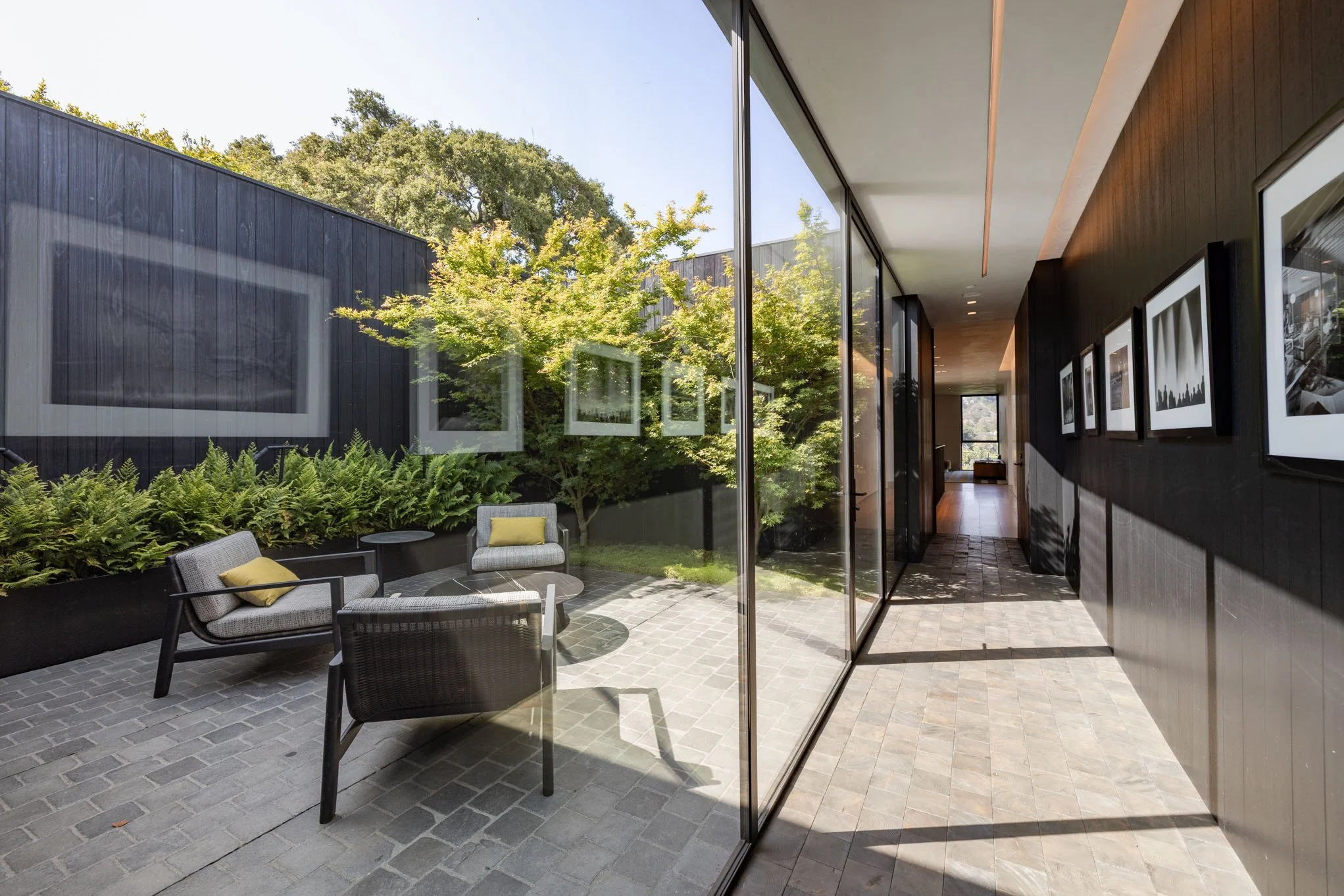 Modern house with a glass wall overlooking a patio with outdoor seating and green trees.