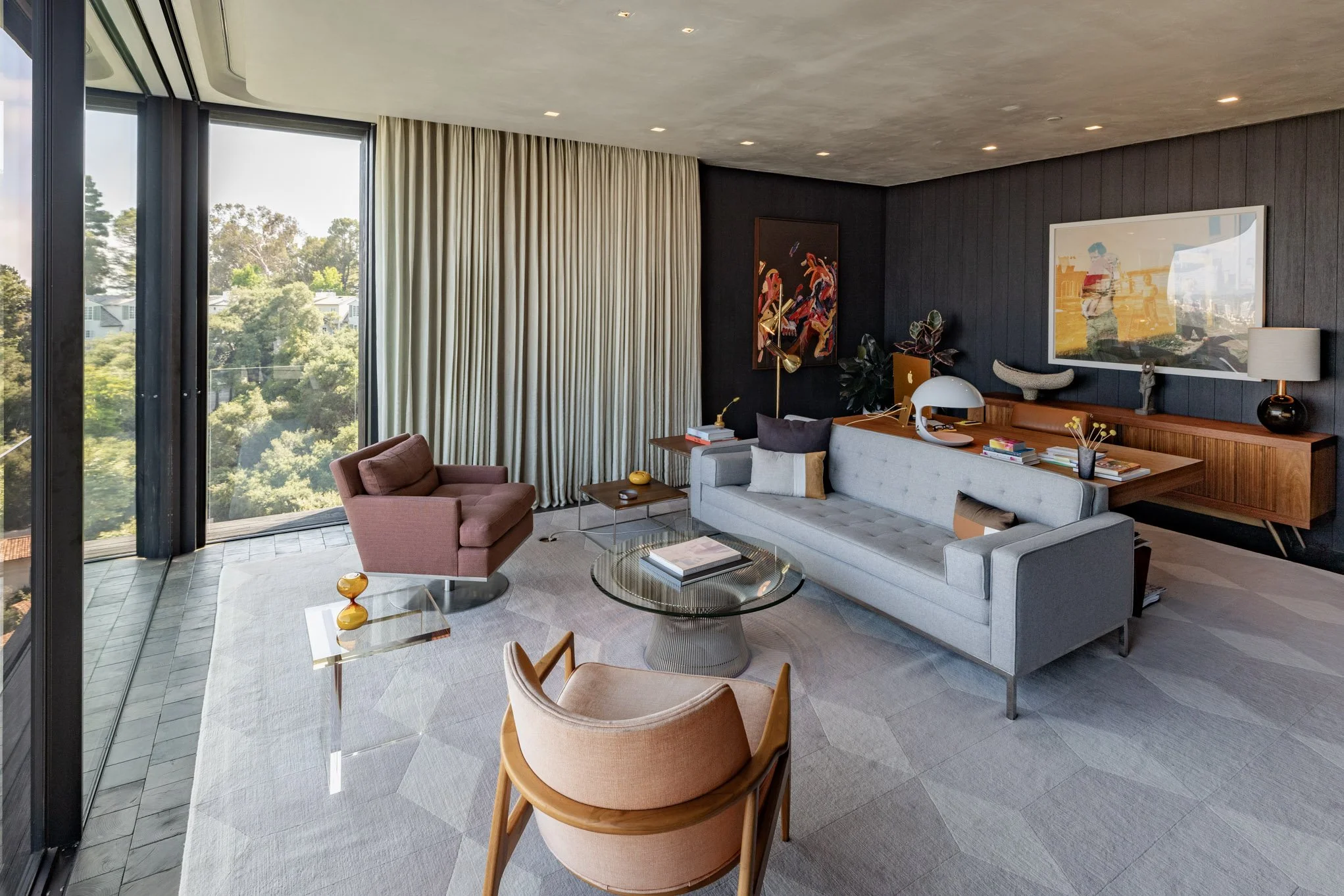 Modern living room with large windows showing trees outside, featuring a gray sofa, pink armchair, beige chair, glass coffee table, side table, wooden sideboard, art wall decor, and decorative lamps.