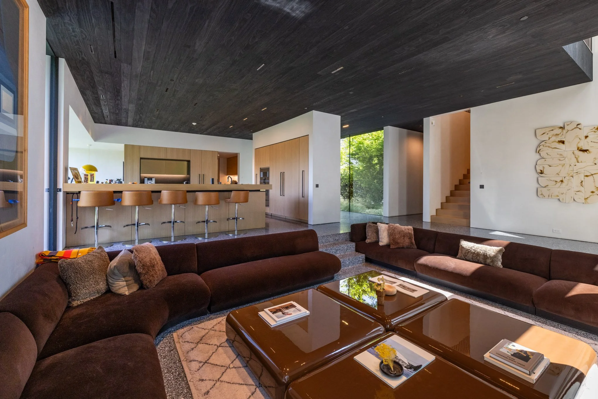 Modern living room with a dark brown upholstery sectional sofa, a wood coffee table, throw pillows, and a rug, with a kitchen area in the background featuring a wooden counter and barstools, and large windows revealing greenery outside.