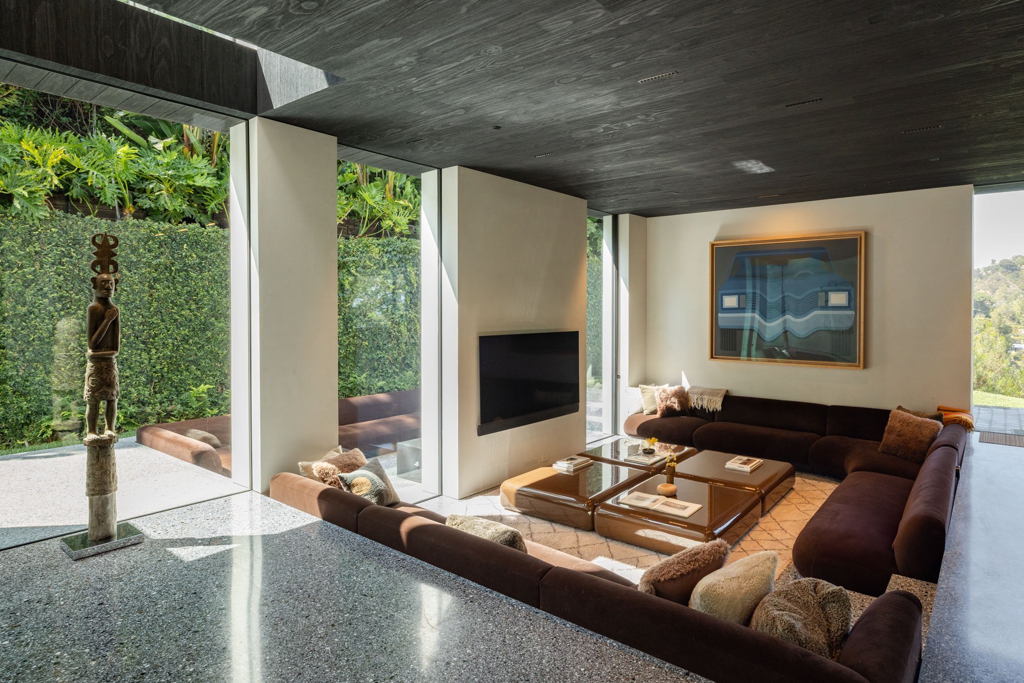 Modern living room with large windows, brown sectional sofa with cushions, abstract art on the wall, and a flat-screen TV.