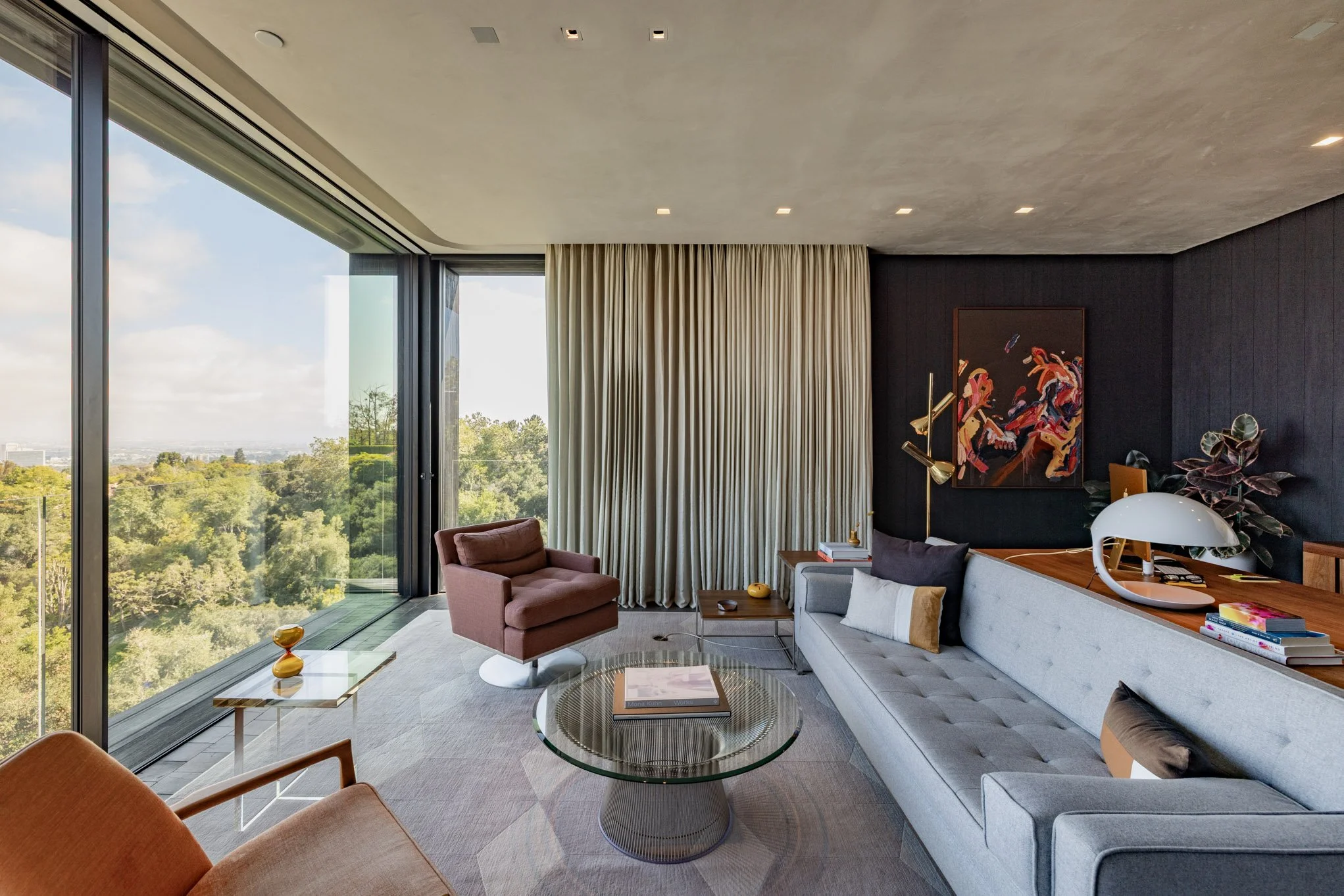 Modern living room with large floor-to-ceiling windows overlooking greenery, beige curtains, a gray sofa with pillows, a brown armchair, glass coffee table, and artwork on the dark accent wall.