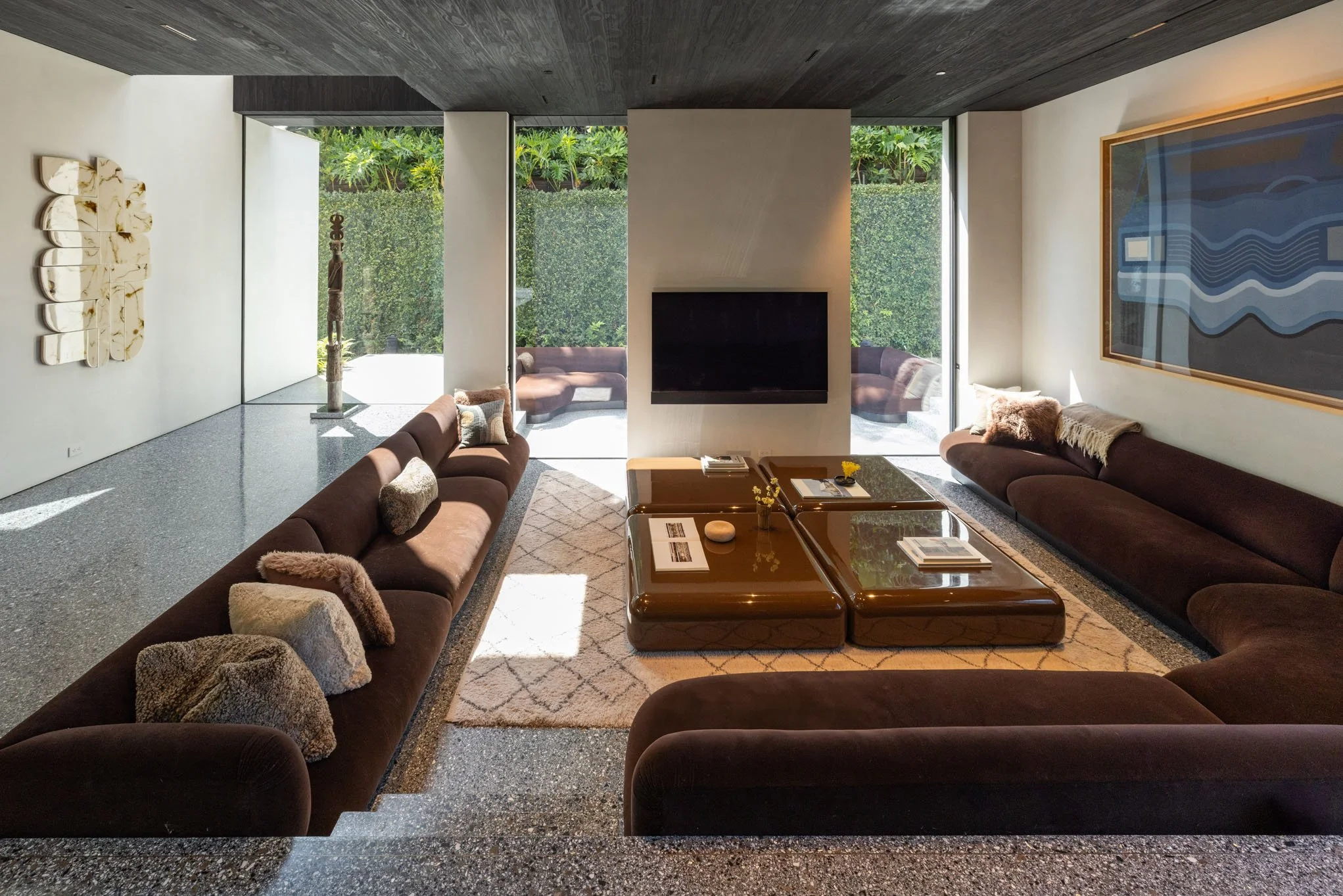 Modern living room with brown sofas, glass coffee tables, a wall-mounted TV, large windows with view of a green garden, abstract art on the walls, and textured carpeting.