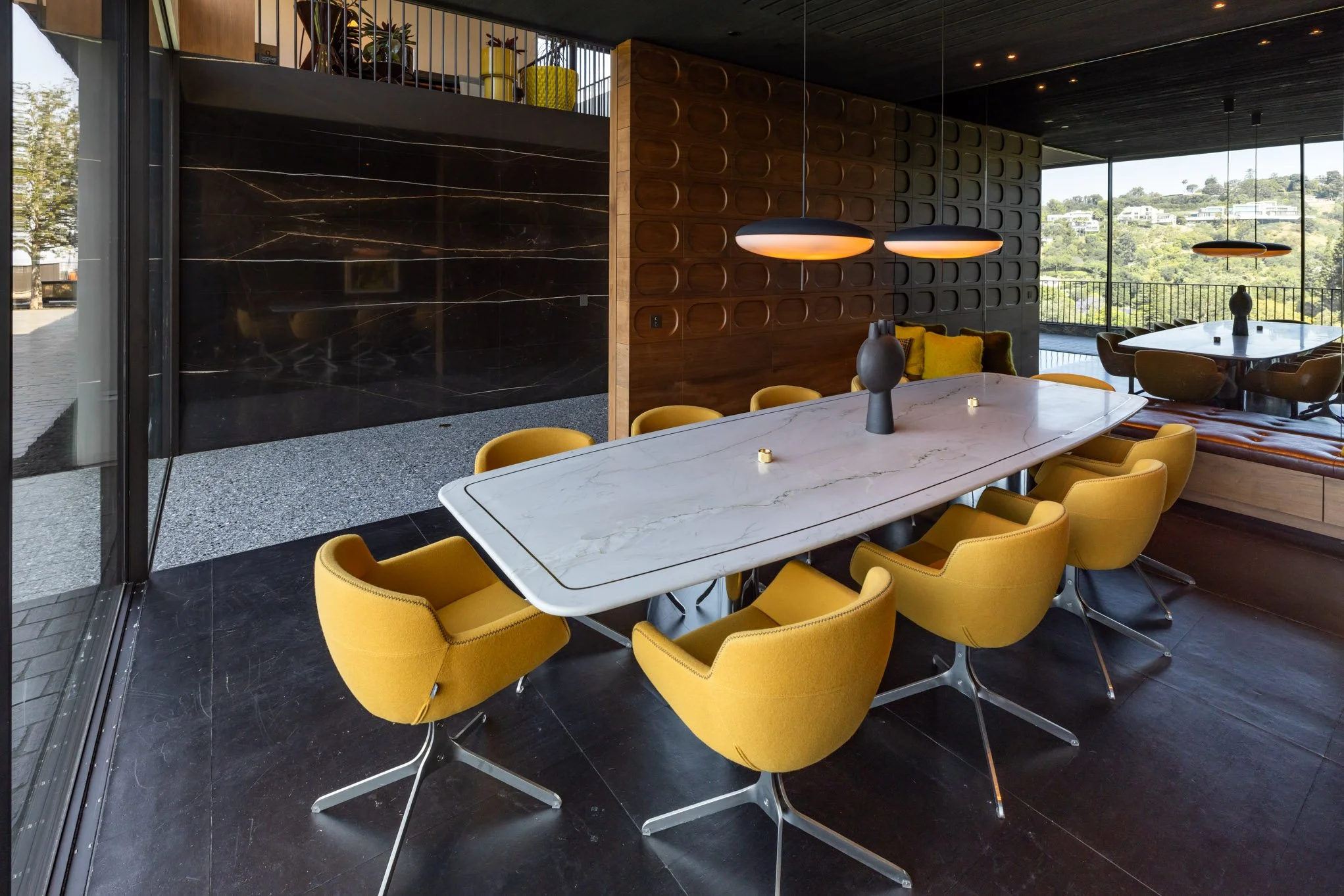 Modern dining room with a large white marble table surrounded by yellow chairs, black pendant lights above, and a view of a hillside landscape through large windows.