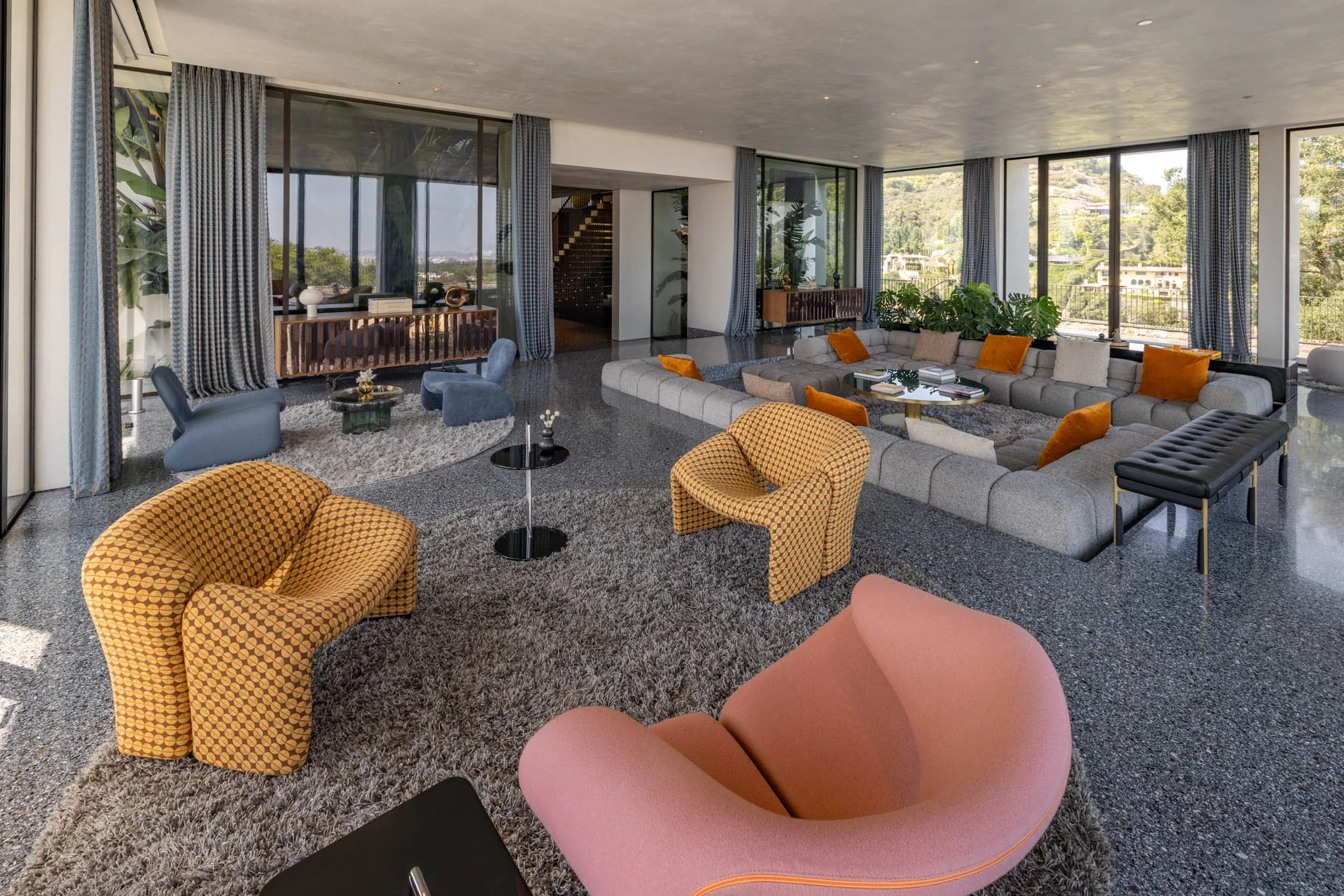 Spacious living room with large windows, gray sectional sofa with orange and beige pillows, pink and yellow accent chairs, round coffee tables, and decorative plants.