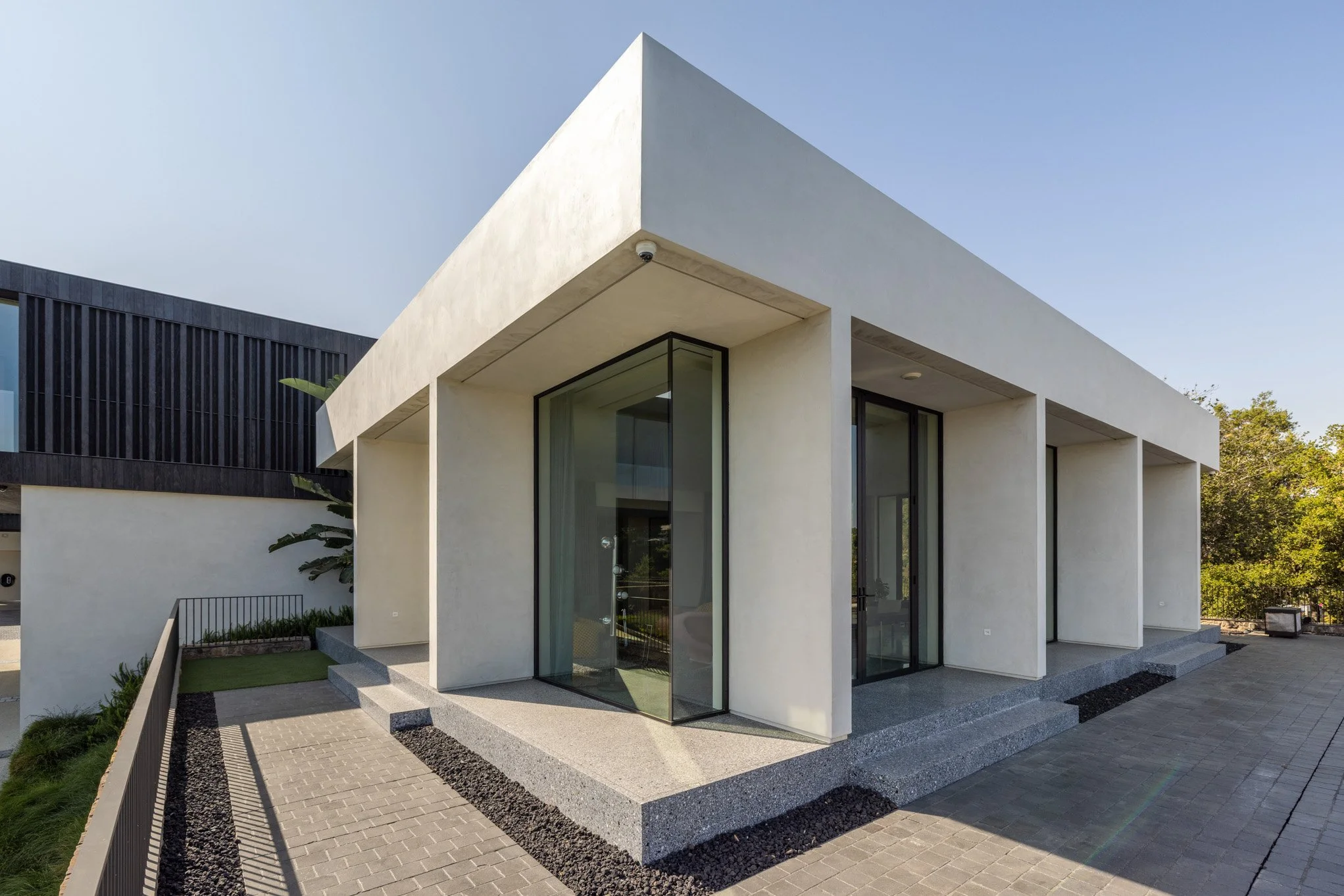 Modern white building with large glass windows and a stone pathway leading to entrance, surrounded by greenery under a clear sky.