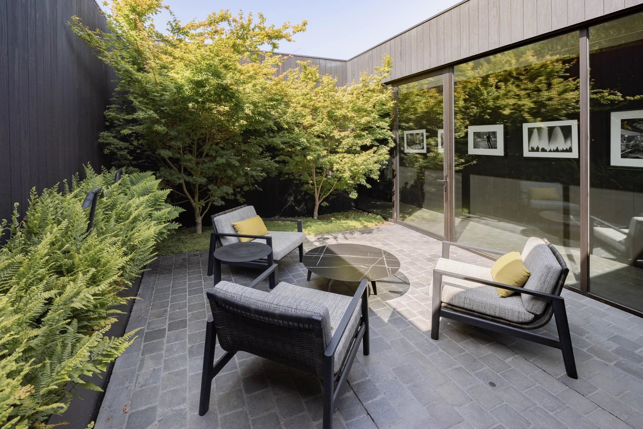 An outdoor patio with four modern chairs around a black marble table, enclosed by a black wooden wall and large glass windows reflecting trees and sky, greenery including ferns and small trees surrounding the patio.