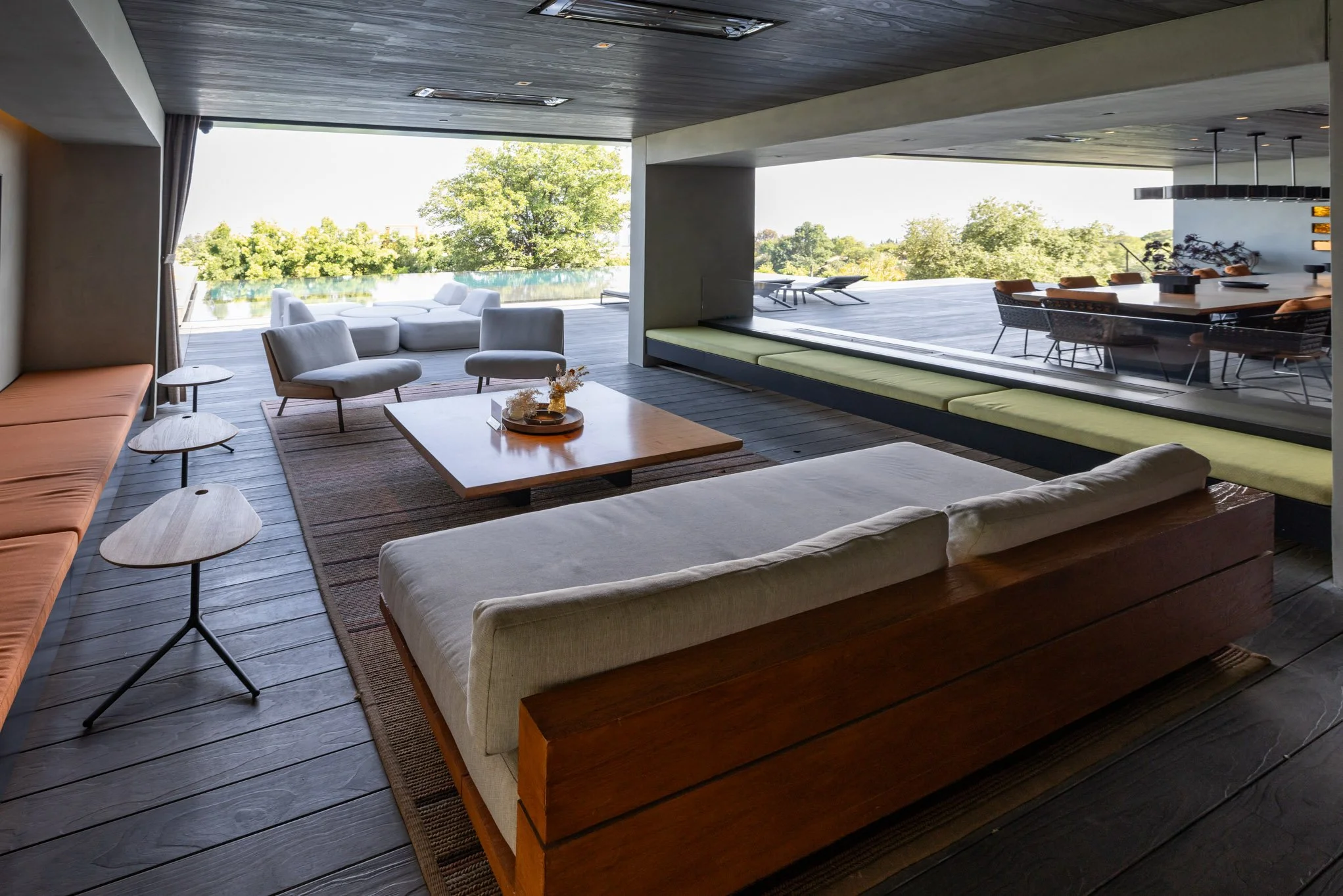 Modern living room with large glass sliding door, offering a view of an outdoor patio and greenery. The interior features a white sofa, light-colored armchairs, a low wooden coffee table with decorative items, small side tables, and a long built-in w