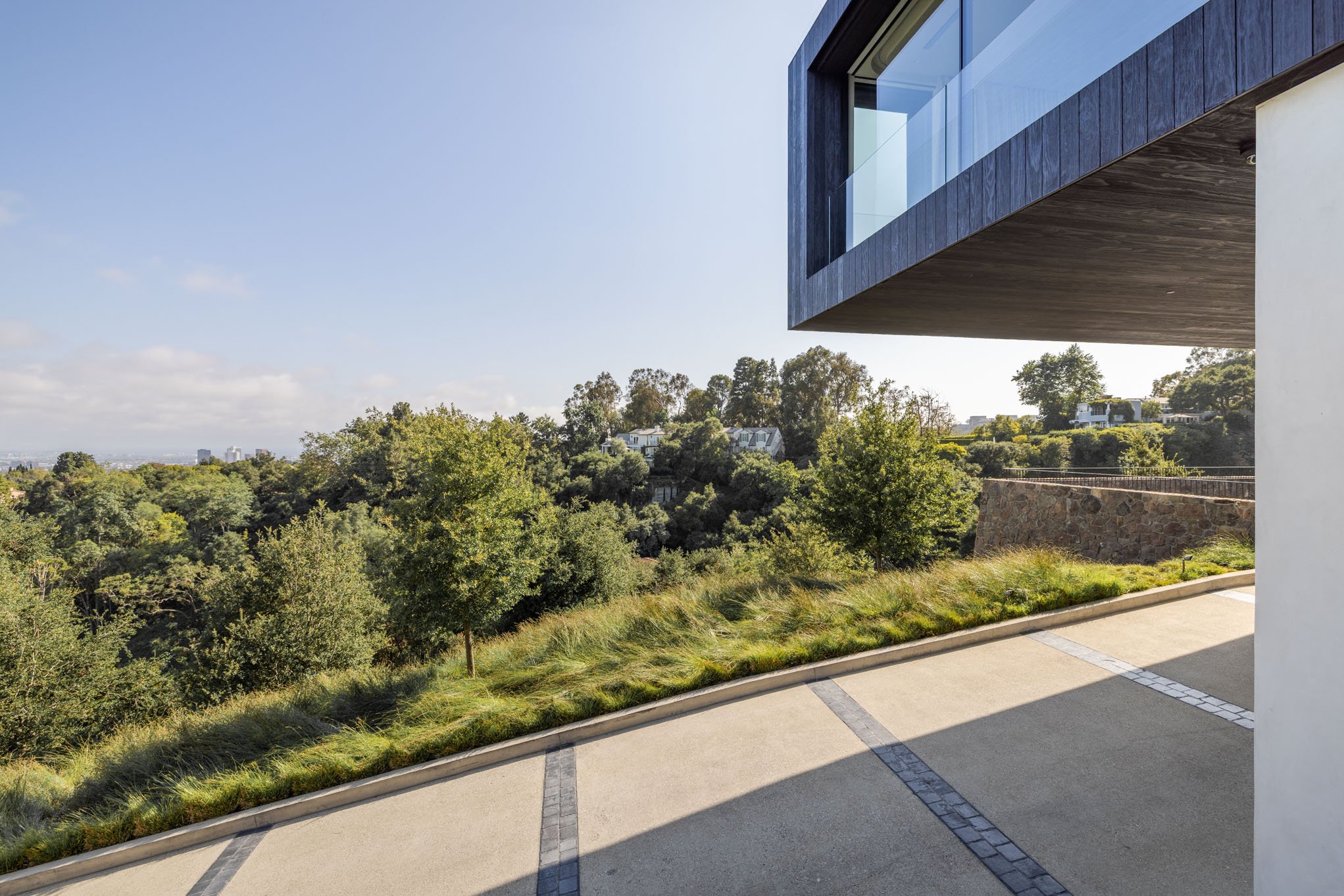 Modern house with a large overhanging balcony, overlooking a lush green landscape with trees and houses in the distance on a clear day.