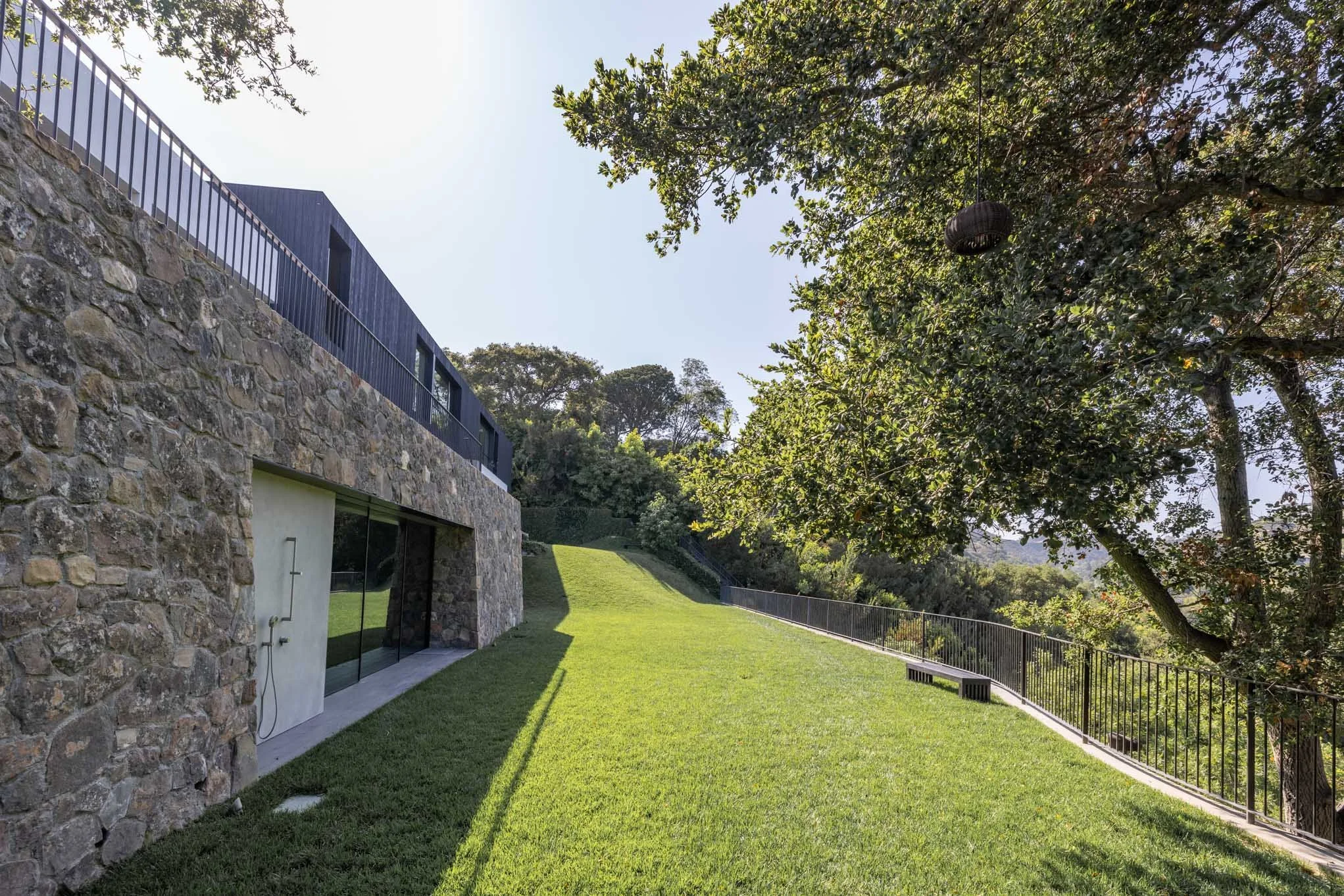 Modern house with stone and black exterior, large glass doors, a spacious grassy yard, trees, and a metal railing on a bright sunny day.