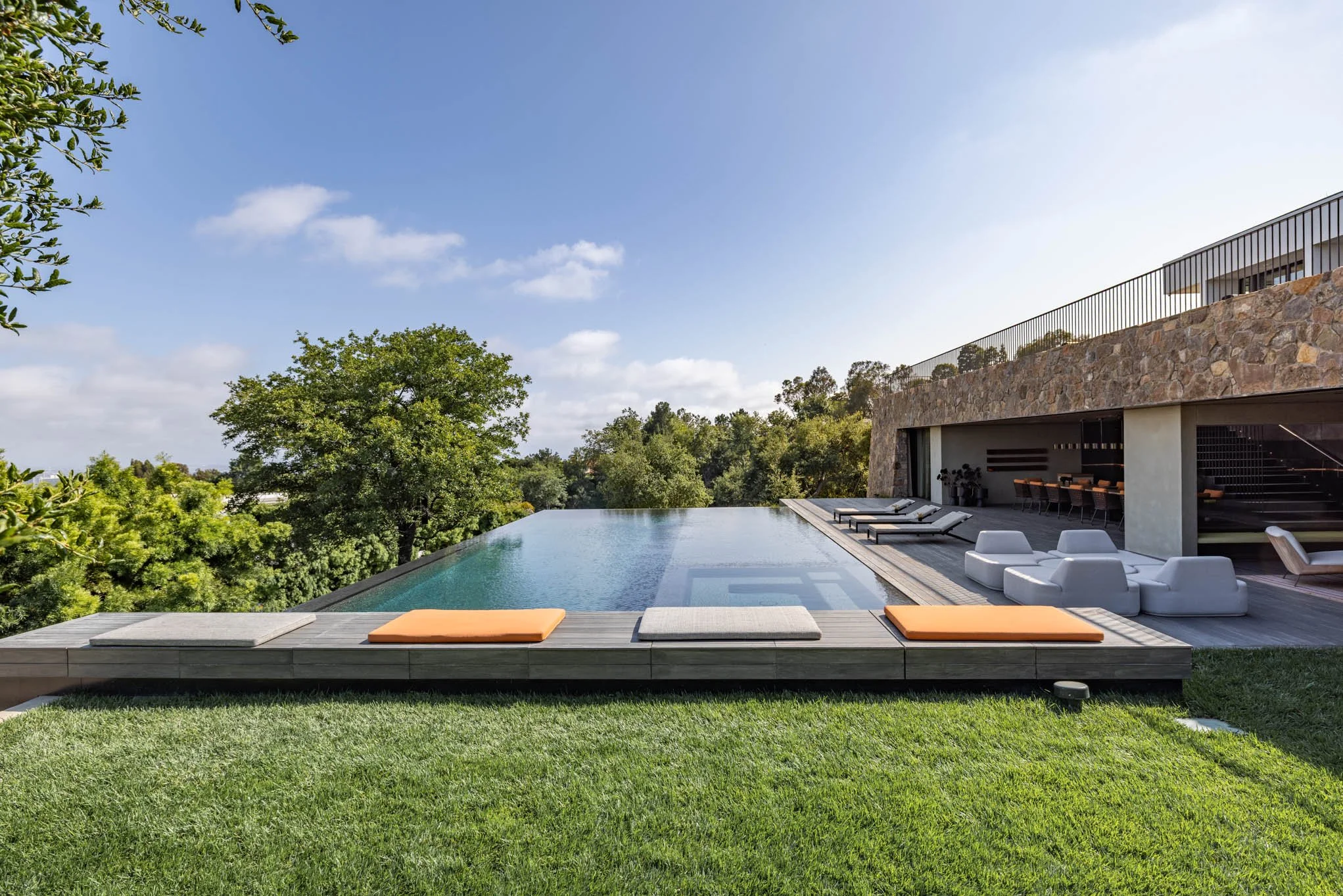 Luxury modern residential property with an infinity swimming pool, lounge chairs, and outdoor seating area, overlooking a lush green natural landscape under a clear blue sky.