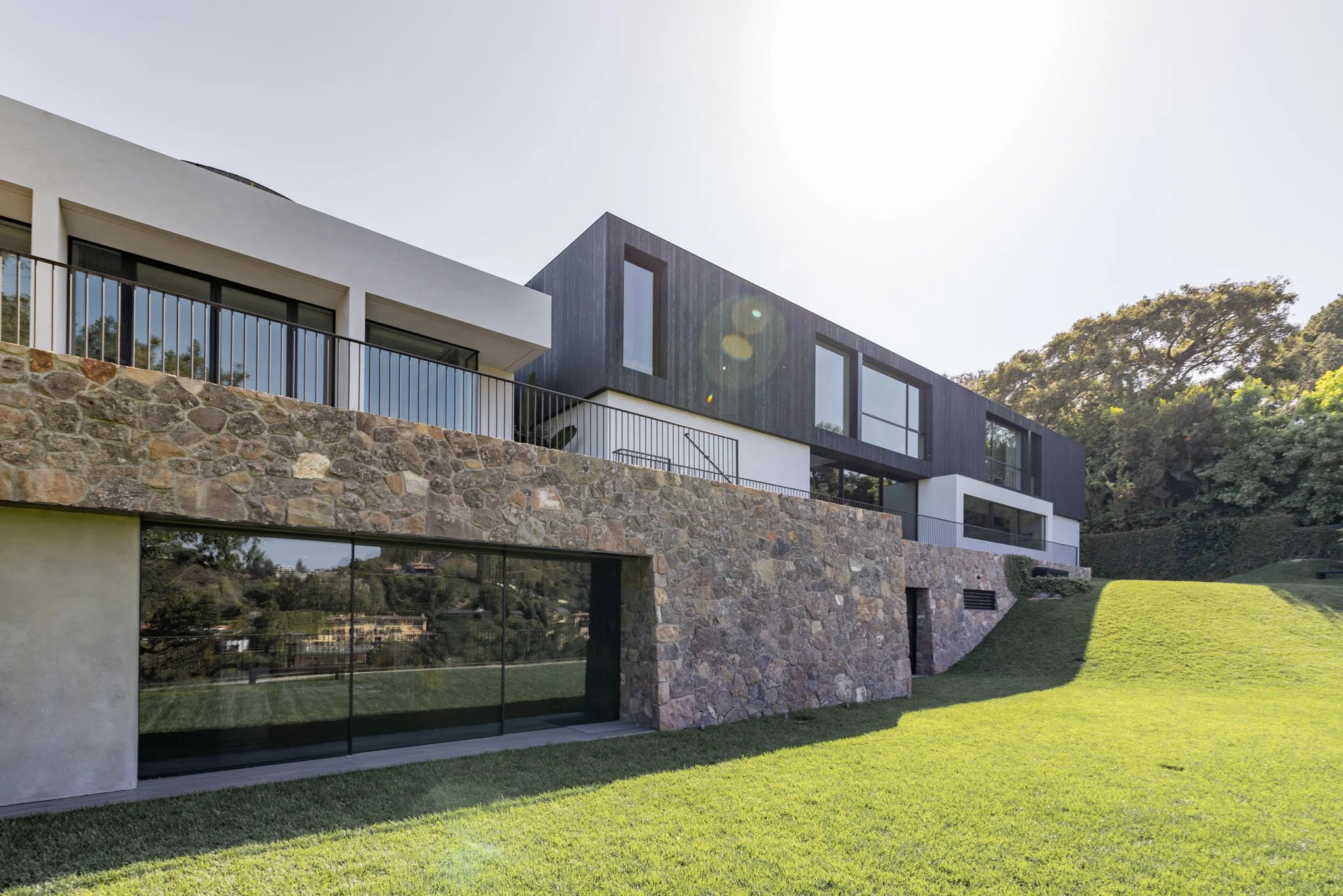 Modern multi-story house with stone, white, and black exterior walls, large windows, and a green lawn in the foreground, surrounded by trees.
