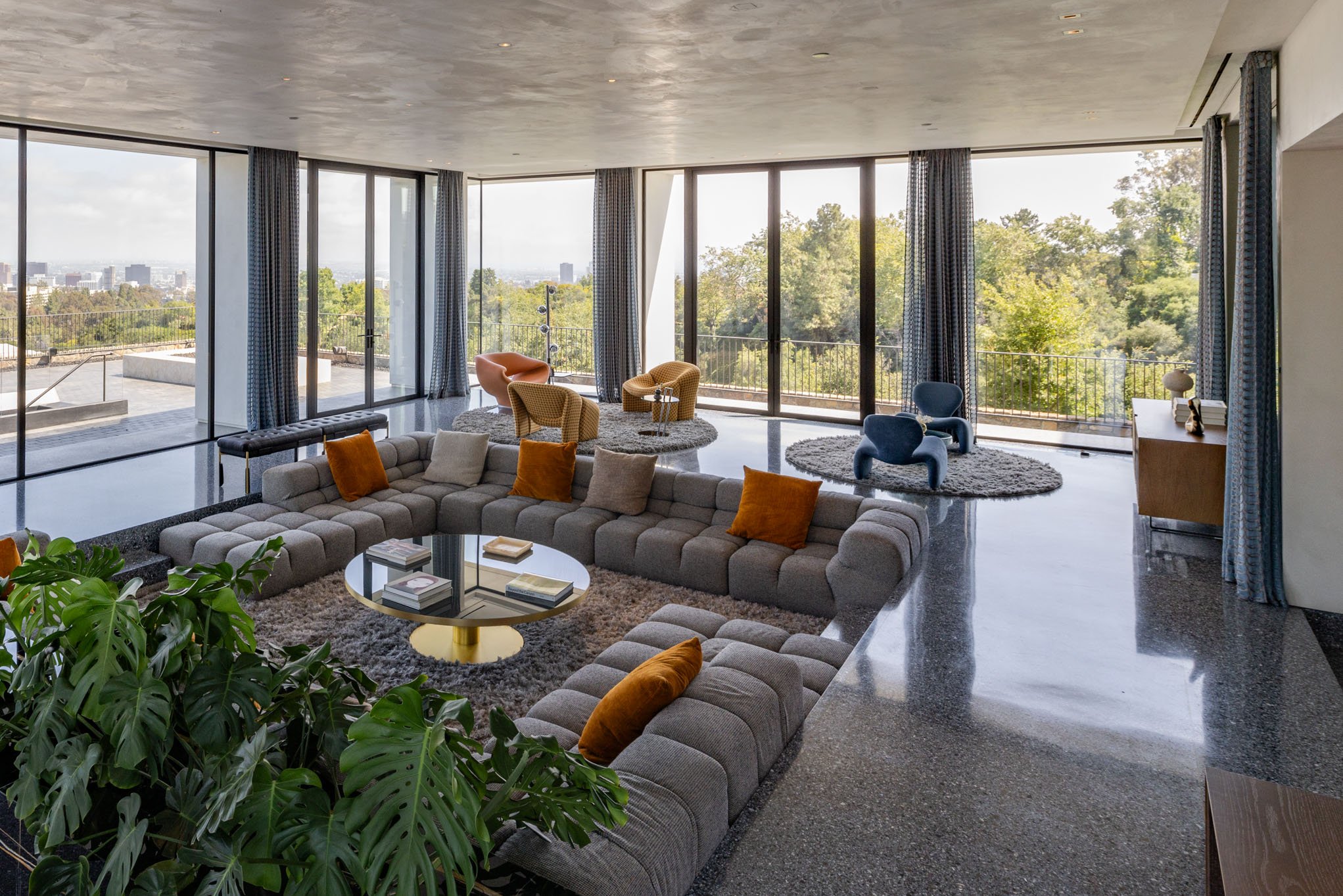 Modern living room with large floor-to-ceiling windows, a large gray sectional sofa with orange and gray pillows, a small black and gold circular coffee table with books, a textured plant in the corner, and various colorful armchairs on round rugs, w